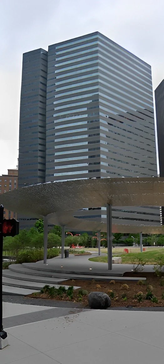 A modern office building with a patterned, striped exterior and a reflective glass facade. In the foreground, there is a concrete structure with supporting poles, a small landscaped garden, and a traffic light showing red.