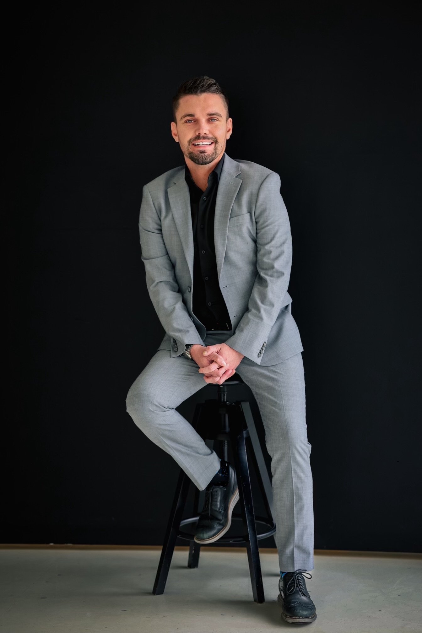 A man in a light gray suit with a black shirt sitting on a tall stool against a black background, smiling, with his hands clasped in front of him.