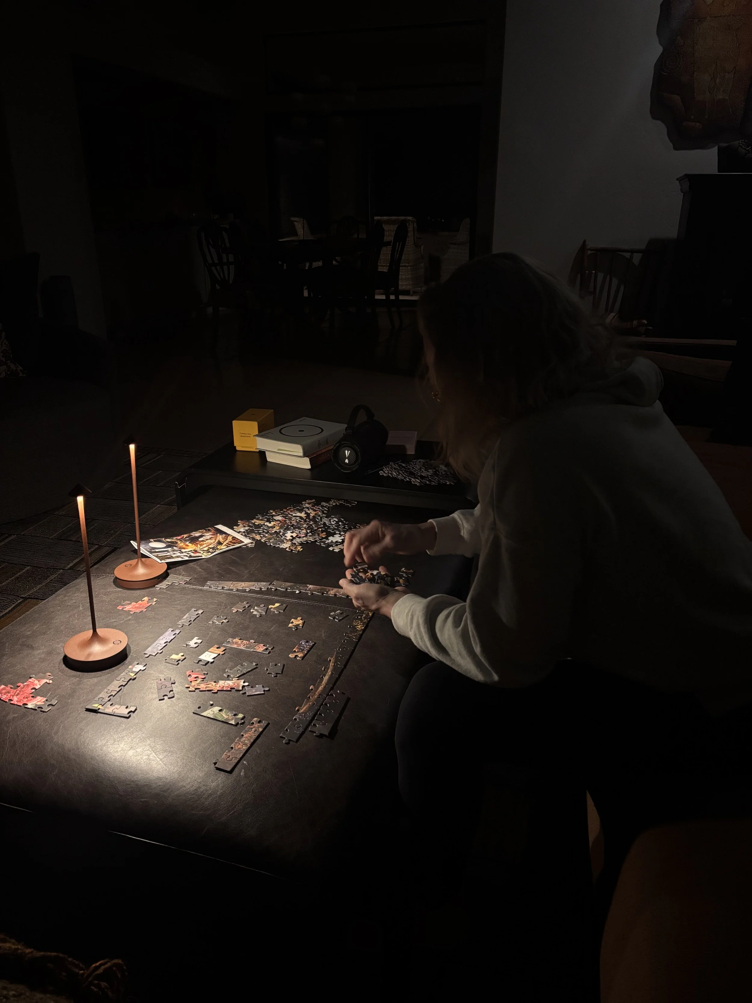Person assembling a jigsaw puzzle on a table in a dimly lit room, with puzzle pieces, a small stack of books, headphones, and candles providing light.