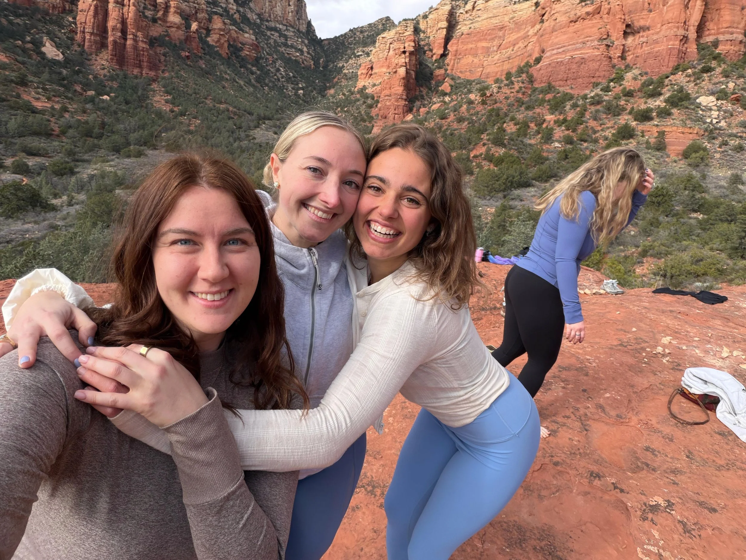 Four women smiling and hugging each other on a red rock formation with a canyon and cliffs in the background, two women are in the foreground and two are in the background, one with blonde hair and another with curly hair.