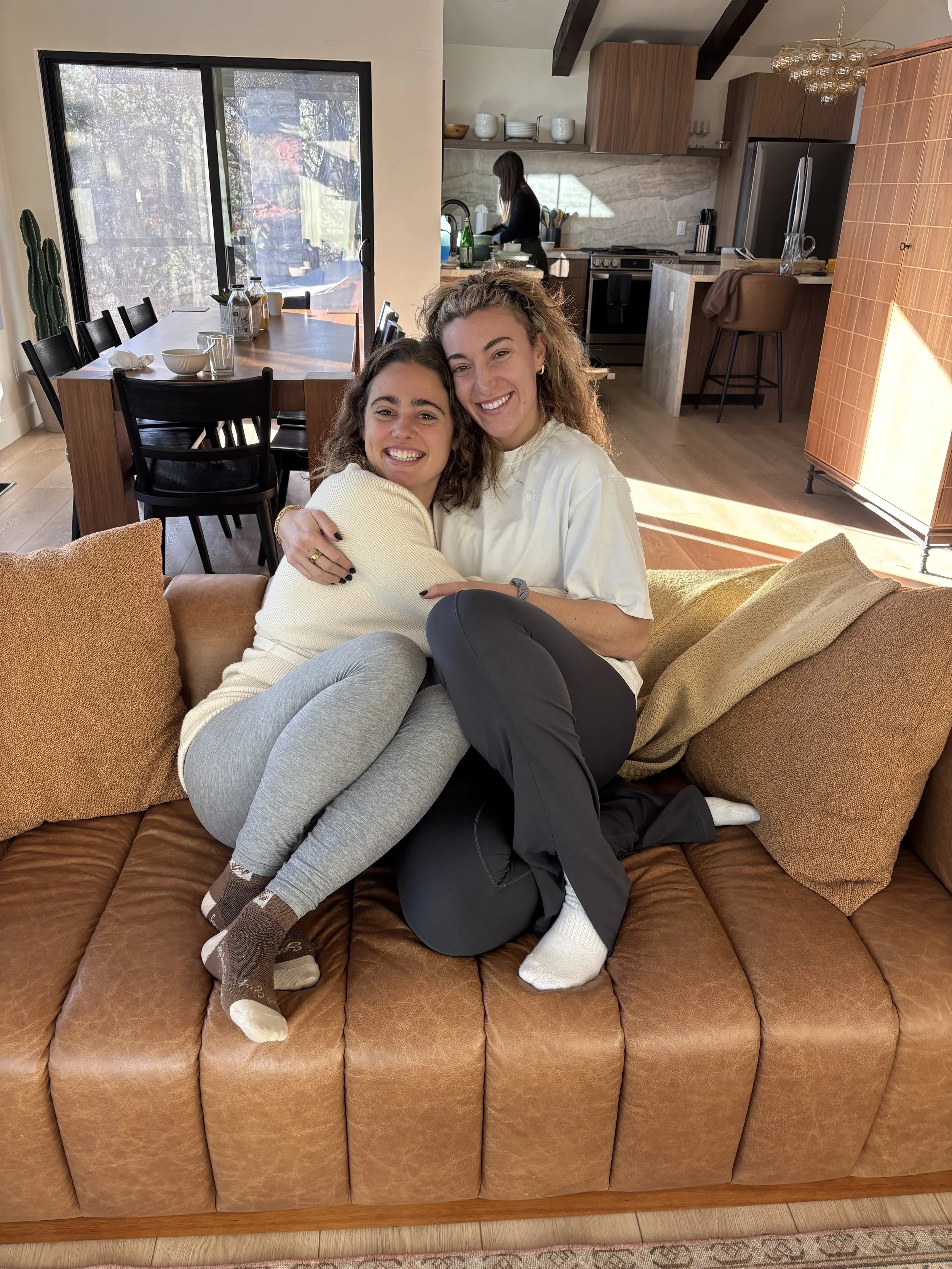 Two women sitting on a brown leather sofa, hugging and smiling at the camera in a cozy living room with a modern kitchen in the background, sunlight streaming in through large windows.