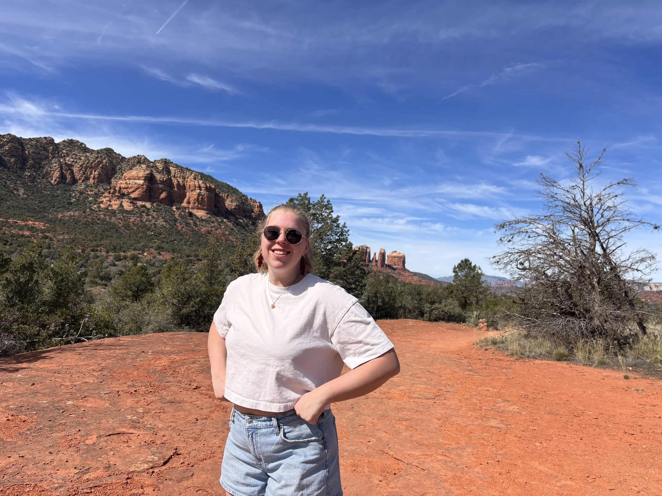 A woman with blonde hair in a ponytail, wearing sunglasses, a white cropped T-shirt, and denim shorts, standing on a red dirt trail in a desert landscape with red rock formations, sparse trees, and a clear blue sky.