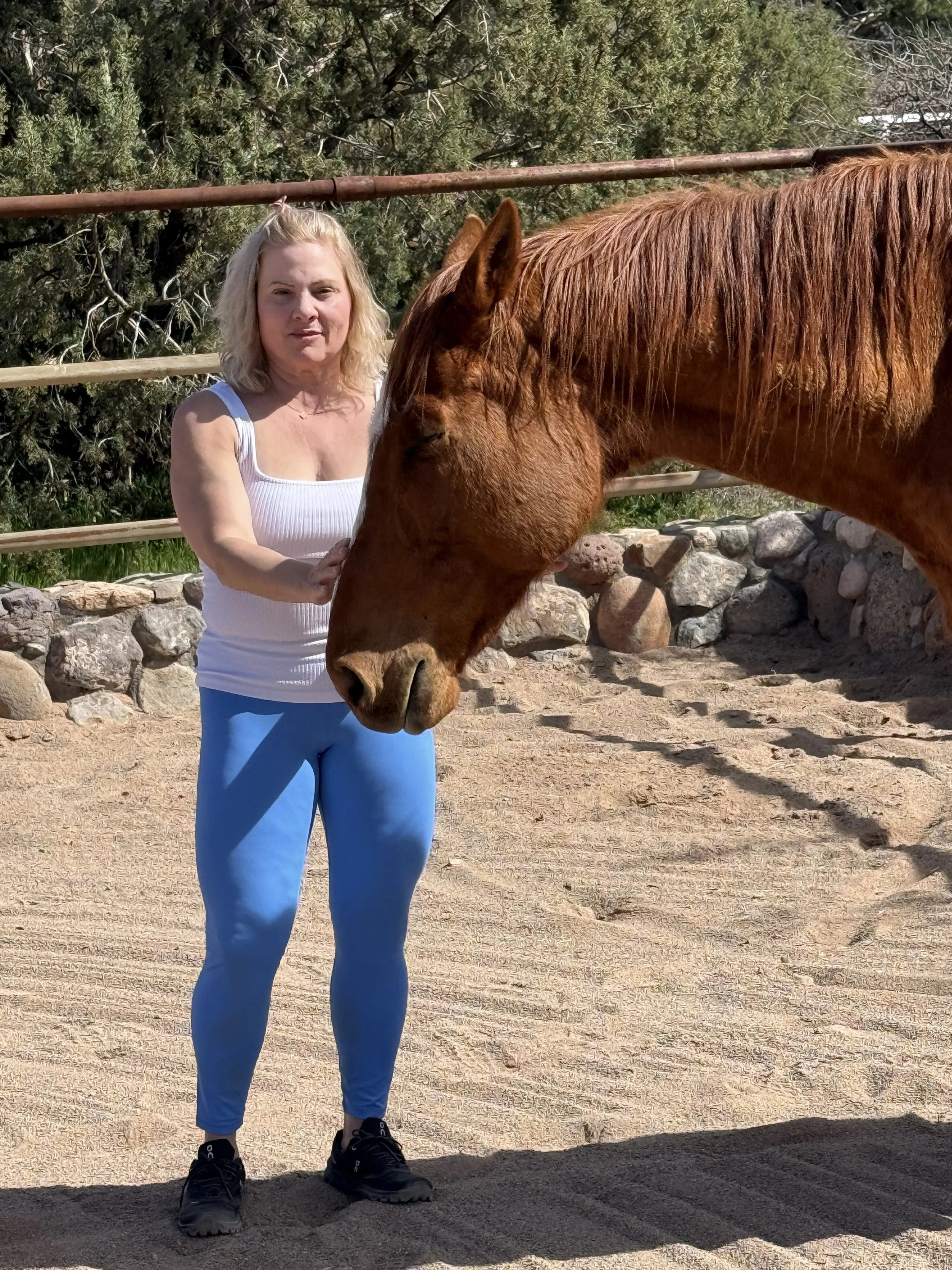 A woman with blonde hair wearing a white tank top and blue leggings standing next to and petting a brown horse in an outdoor sandy area with trees and a stone wall in the background.