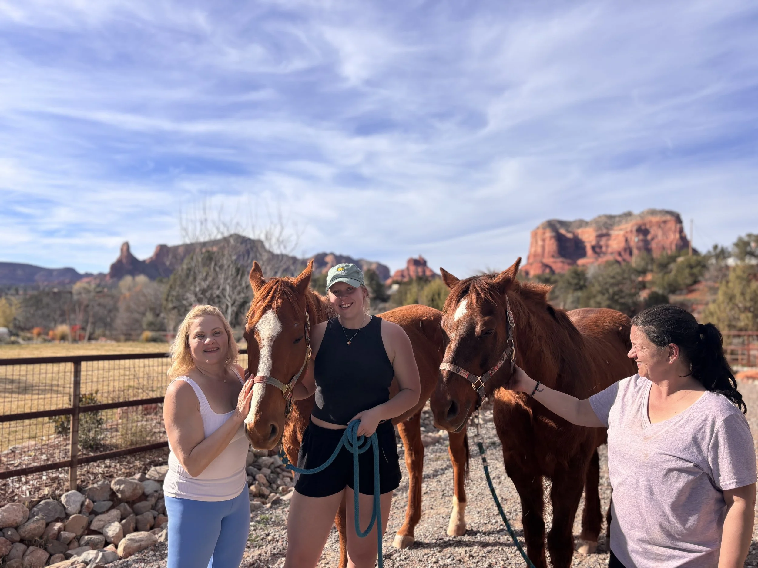 Three women with two horses outdoors, desert mountains in the background, on a sunny day.