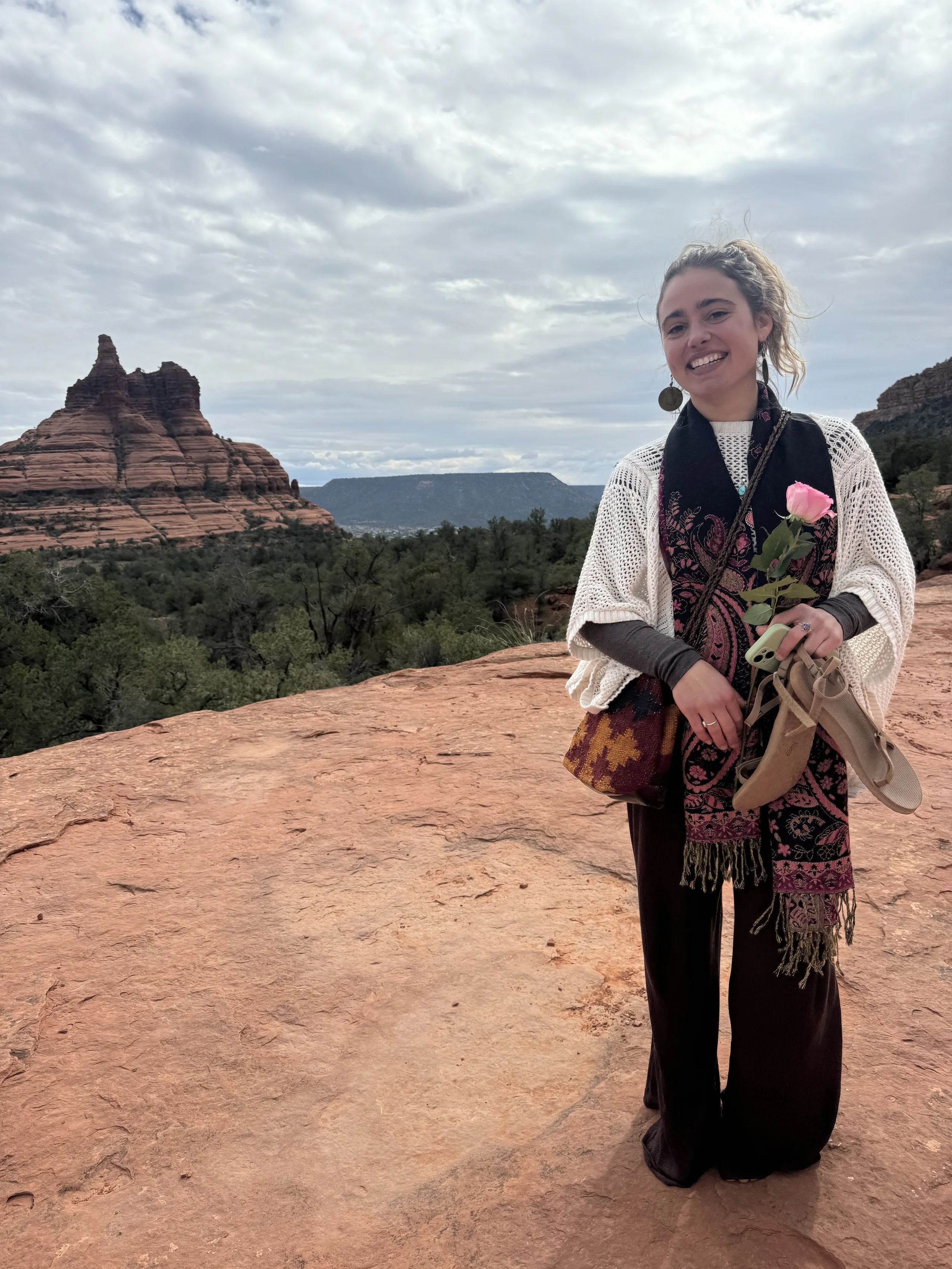 A young woman standing on red rock in a desert landscape, holding a pink flower, with rock formations and cloudy sky in the background.