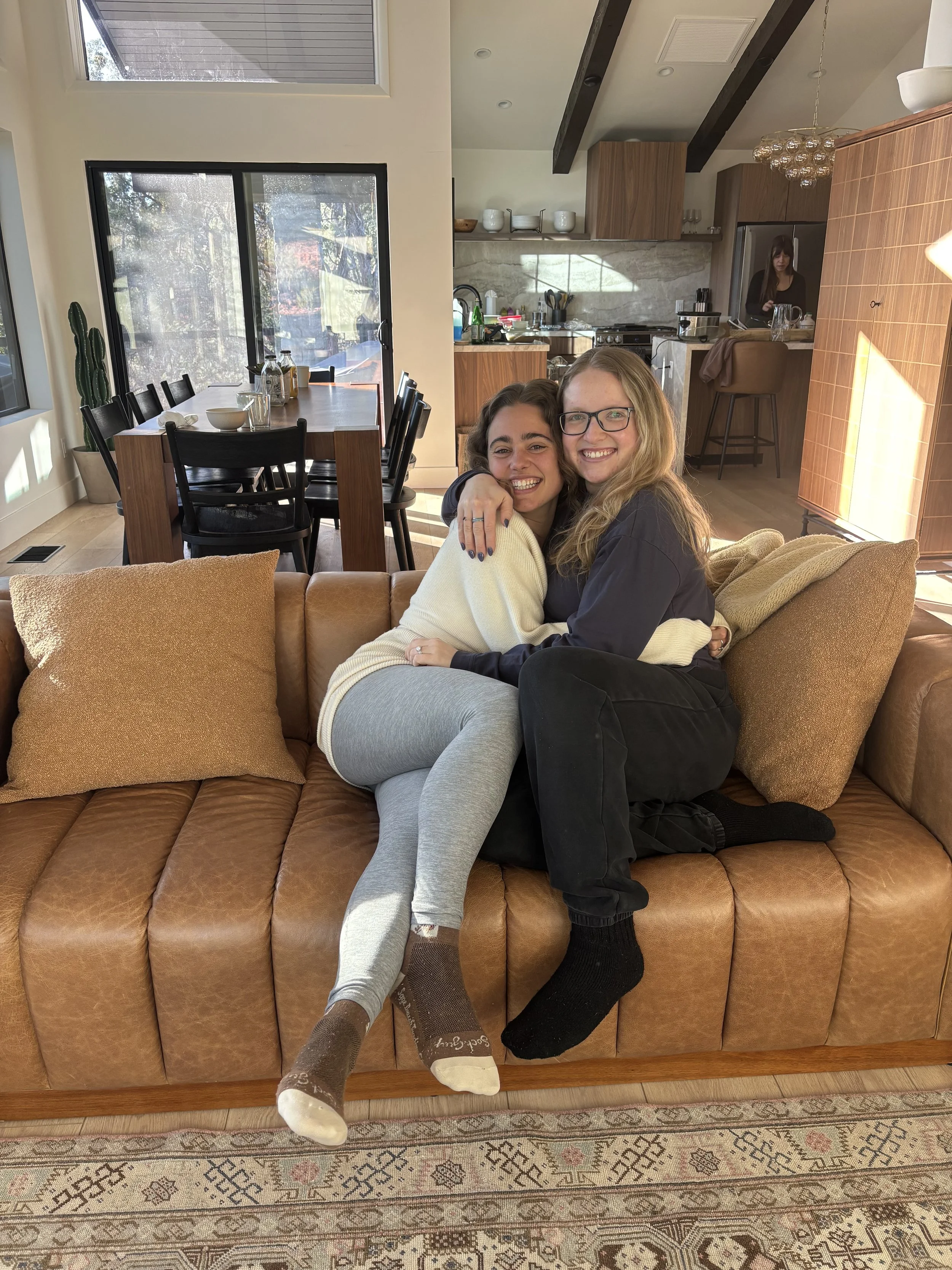 Two women sitting on a brown leather couch hugging and smiling in a cozy living room with a kitchen in the background.
