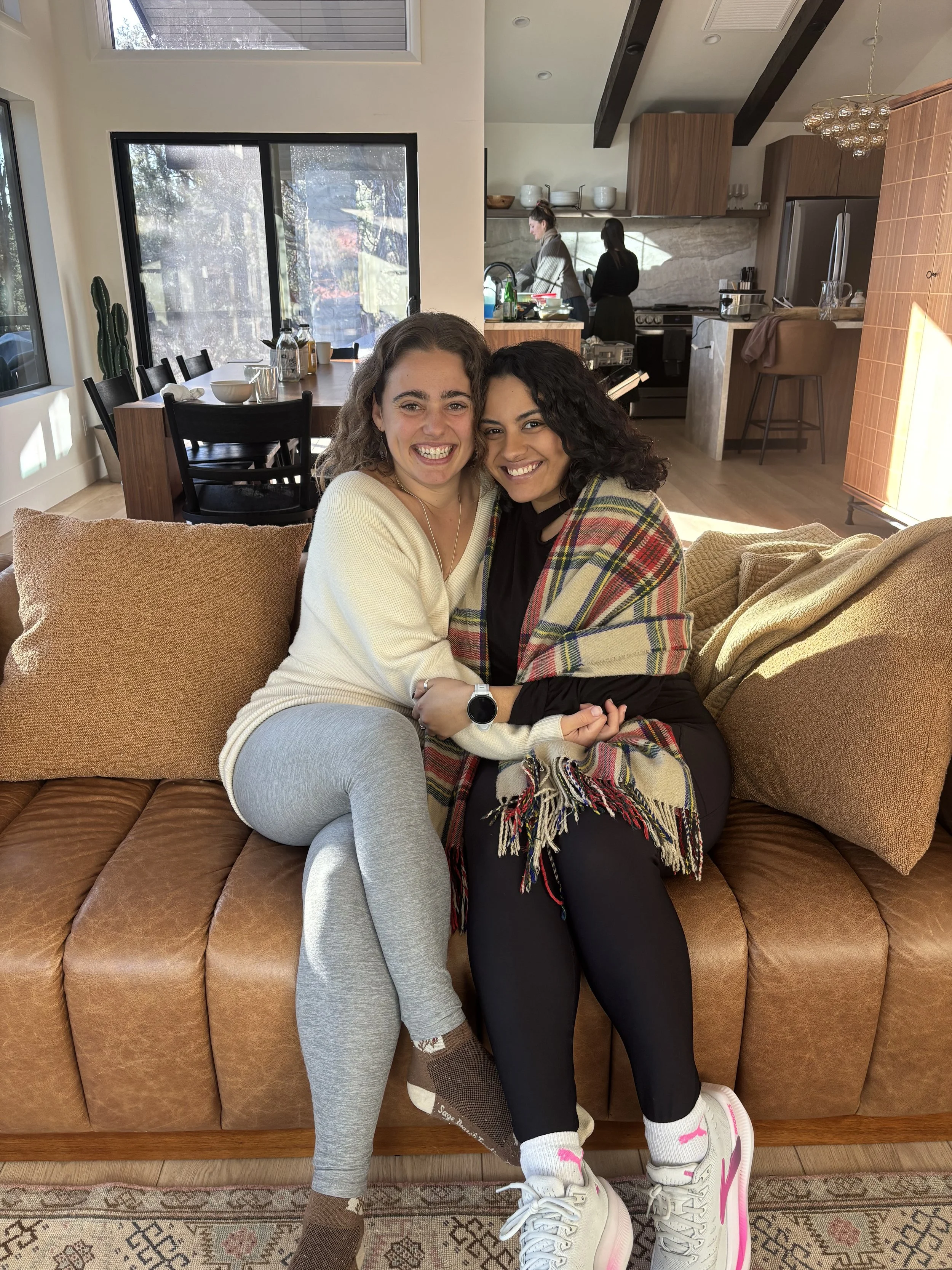 Two women sitting on a brown leather sofa, smiling and embracing each other inside a modern home.
