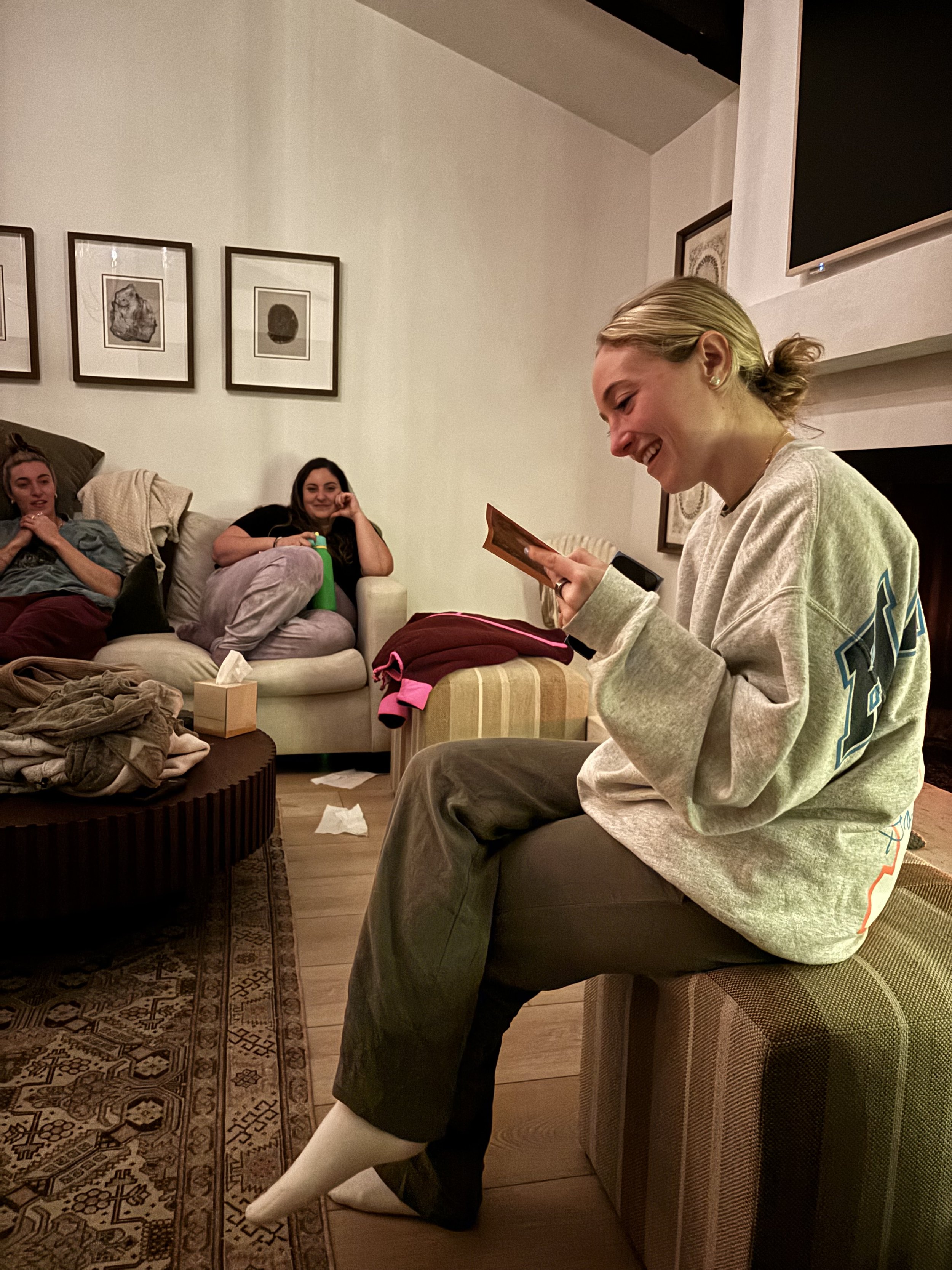 Three women sitting and laughing in a living room, with a woman in the foreground smiling and holding a card or photo. The room has framed artwork on the walls and various items on the coffee table.