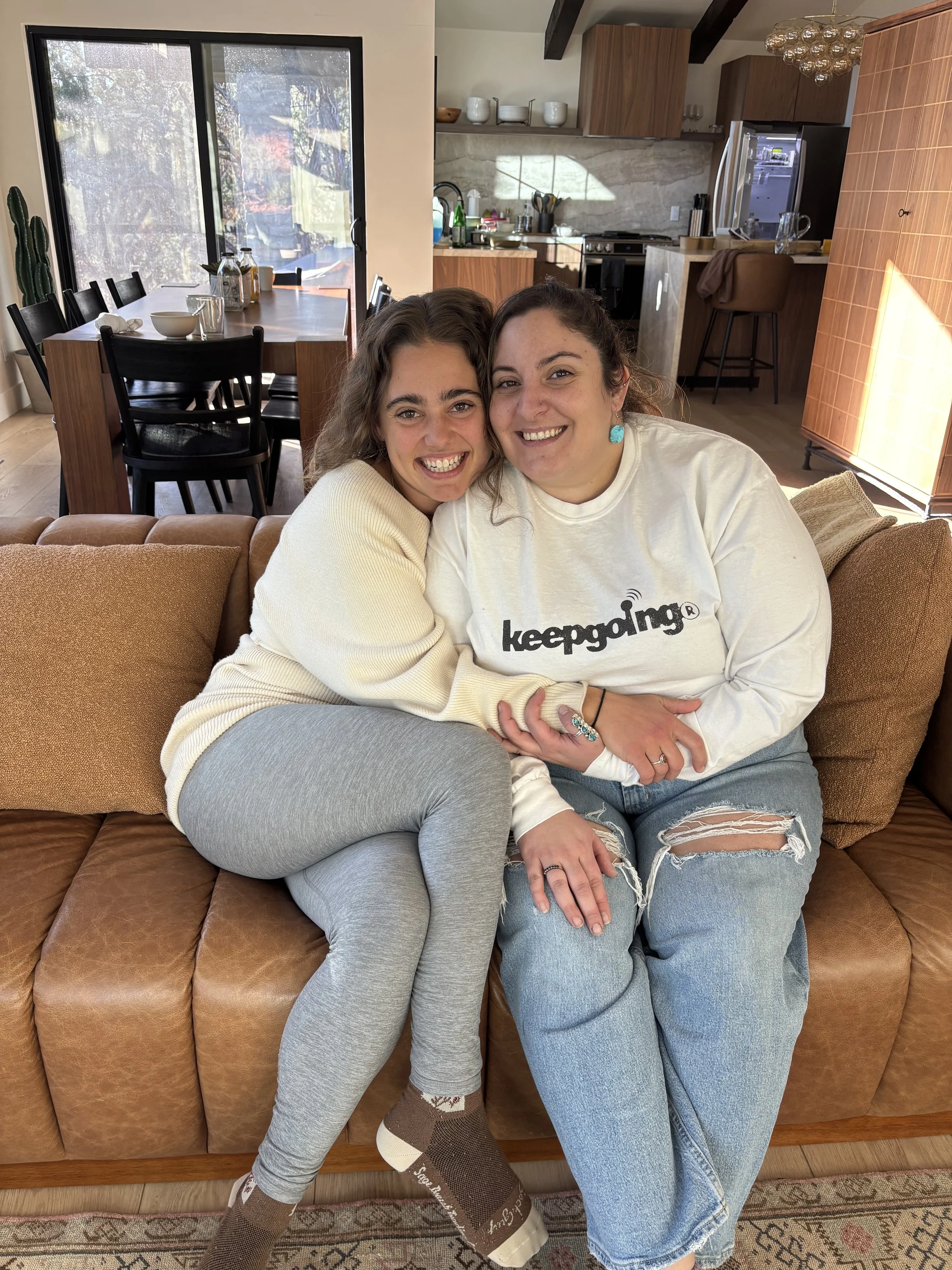 Two women sitting on a brown leather couch, smiling and hugging each other inside a cozy living room with a kitchen in the background.