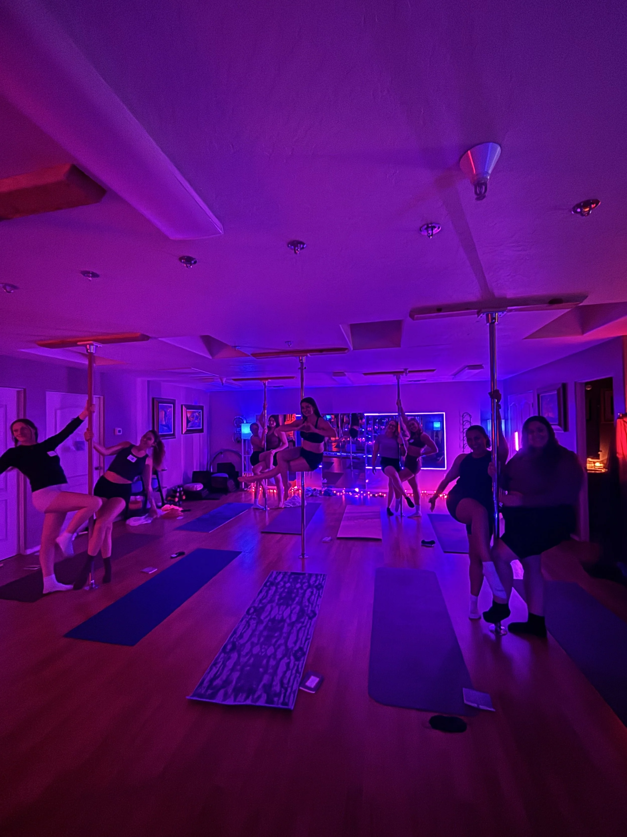 Group of women participating in a pole dancing class in a dimly lit room with purple and blue lighting, exercise mats on the floor, and framed pictures on the walls.