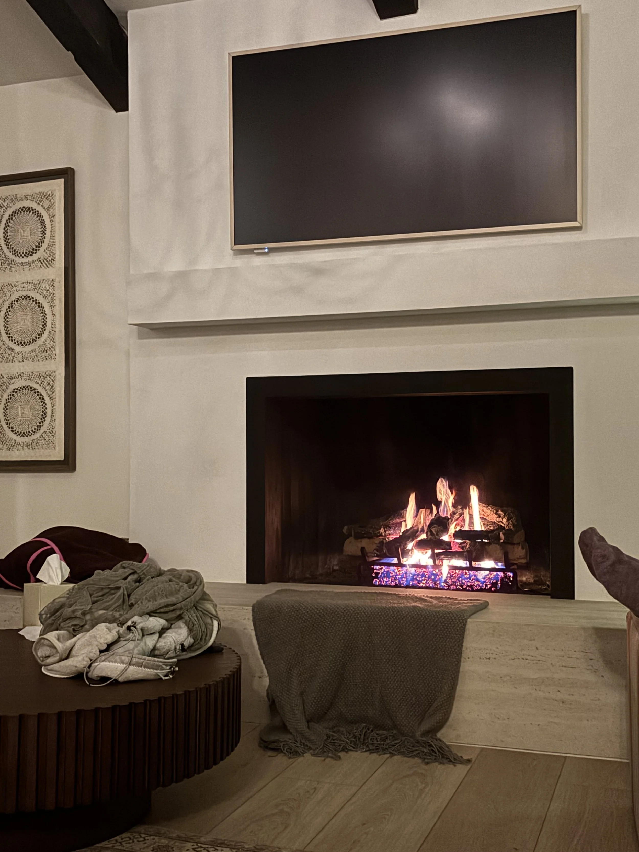 A cozy living room corner with a lit fireplace, a flat-screen TV mounted above, a round wooden table with clothes and a bag, and a beige blanket draped over a small chair or ottoman.