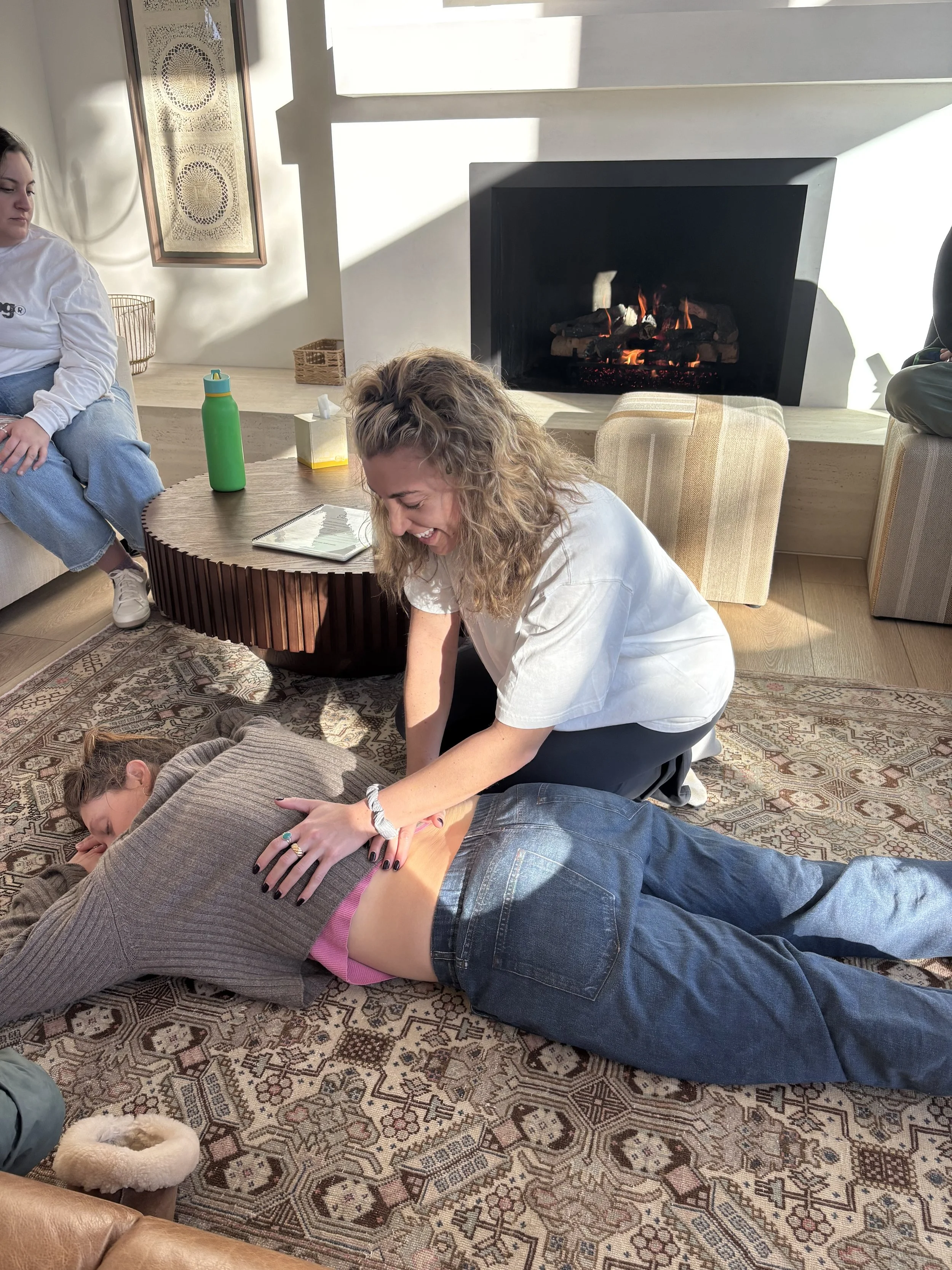 A woman performing an abdominal CPR on another woman lying face down on an ornate rug, with a fireplace and sofas in the background.