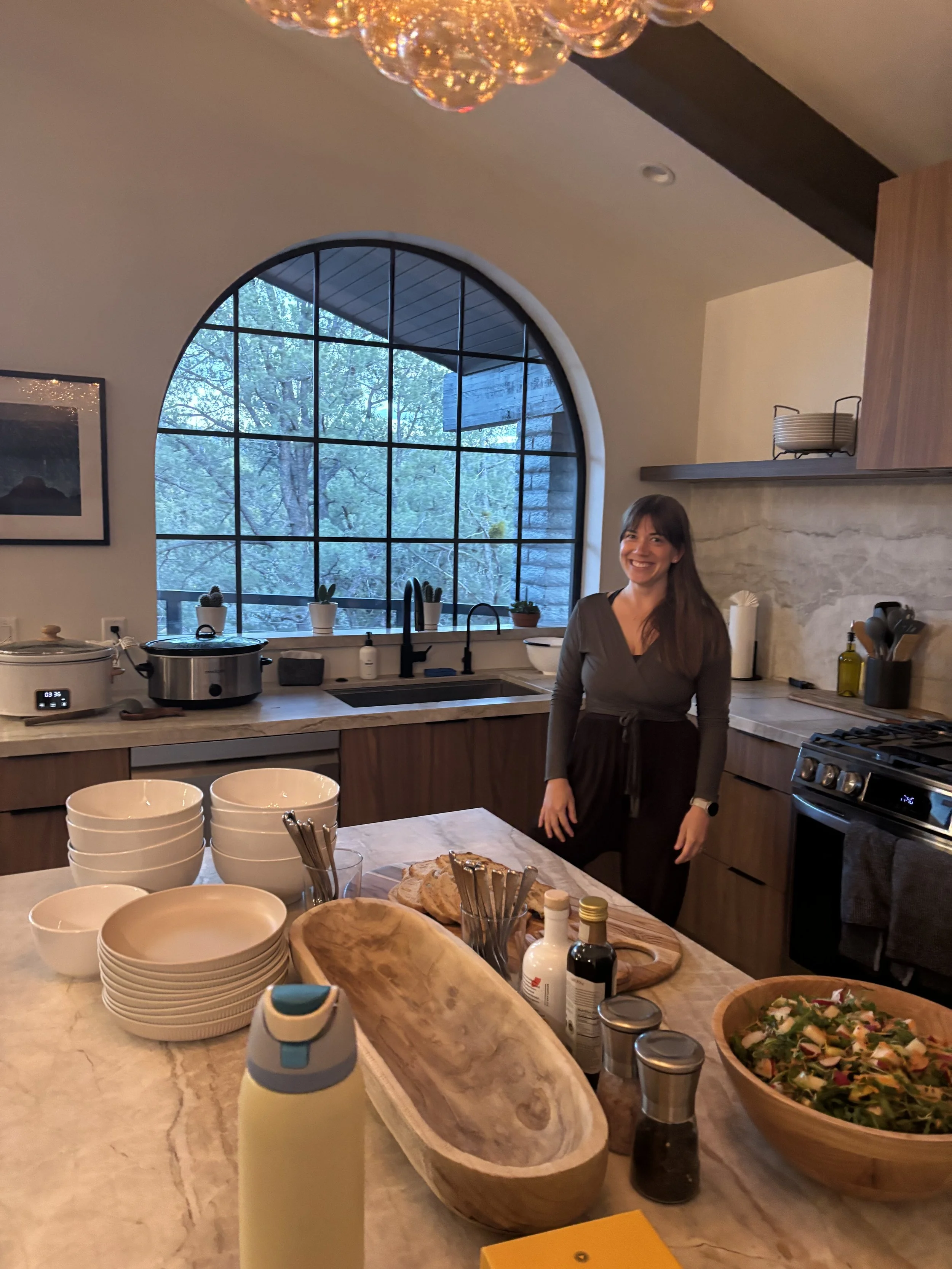 A woman smiling in a kitchen near a countertop with bowls, bottles, and a salad. Large window with trees outside and modern kitchen decor.
