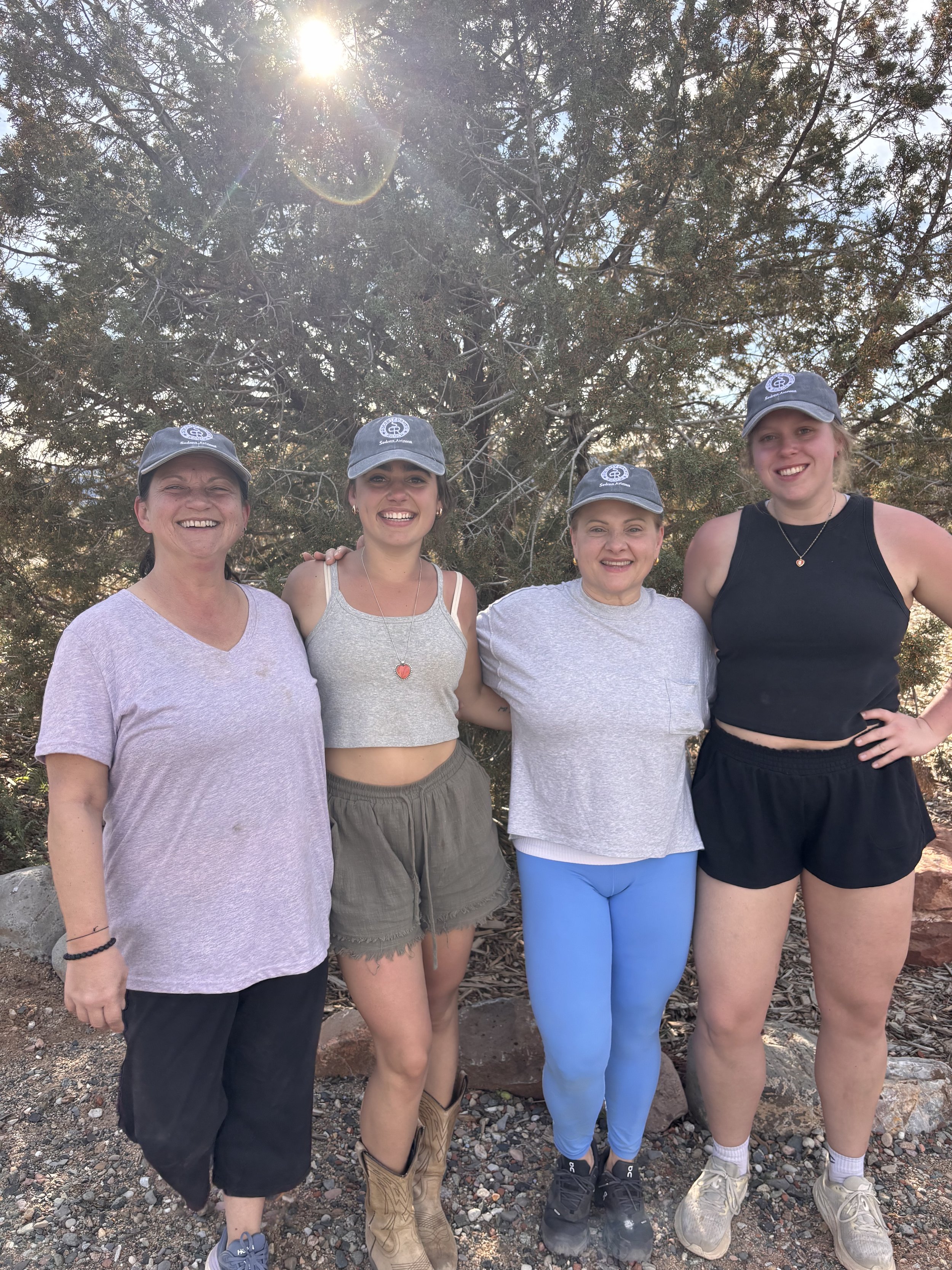 Four women standing outdoors in front of a large tree with sunlight shining down, smiling at the camera, all wearing gray hats with a logo.