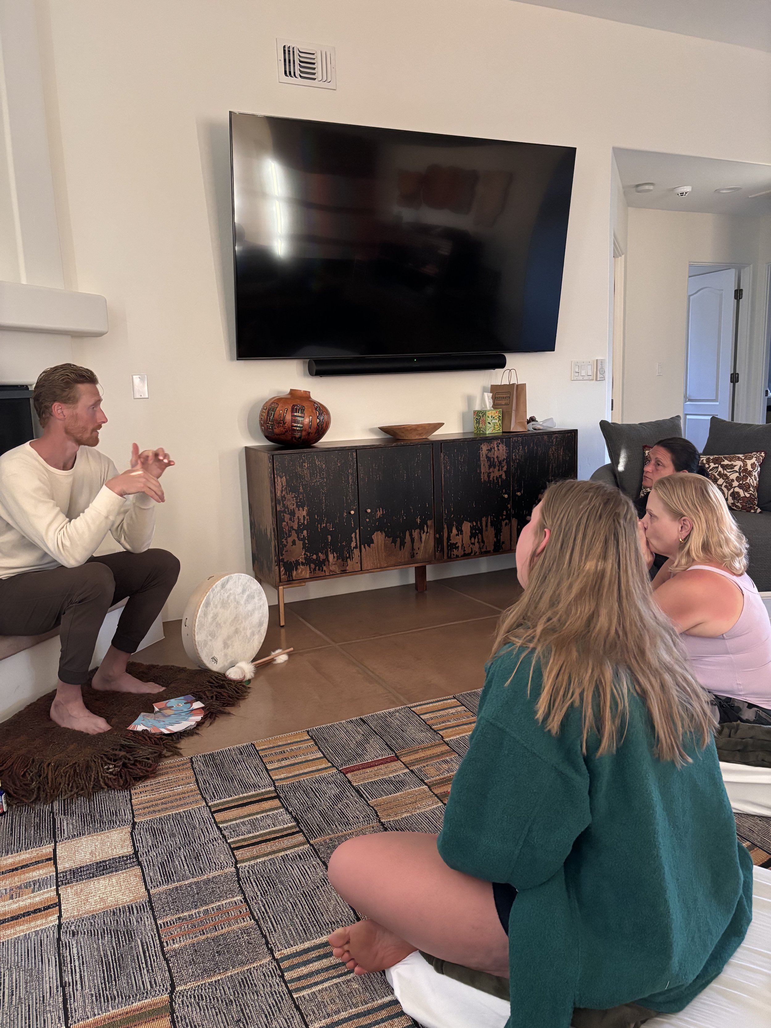 Group of people sitting on the floor watching a man sitting on a ledge; a large television is mounted on the wall in a living room.