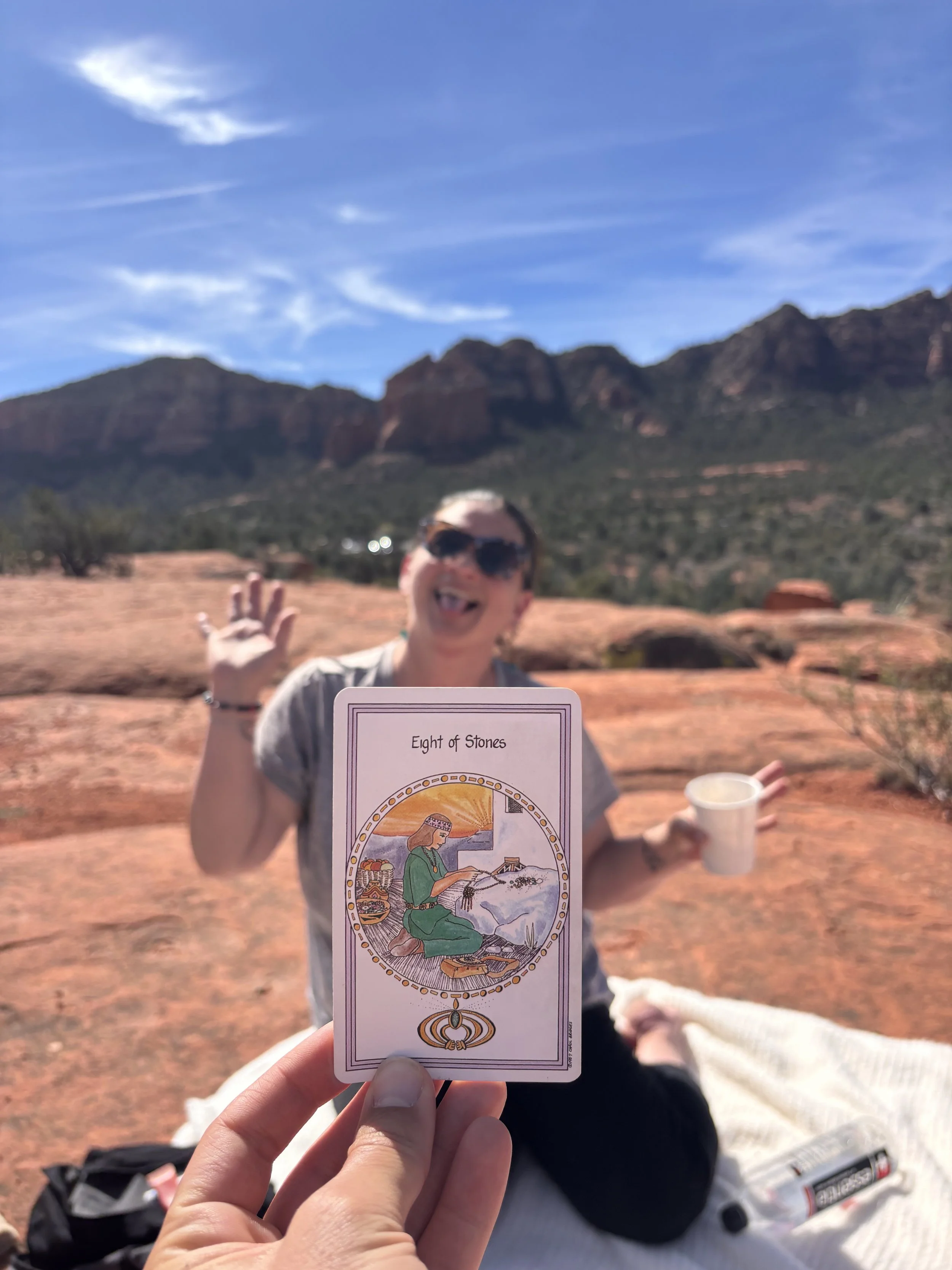 A person holding a tarot card labeled 'Eight of Stones' in focus, with a smiling woman wearing sunglasses and waving, sitting on a blanket in a desert-like landscape with mountains and blue sky in the background.