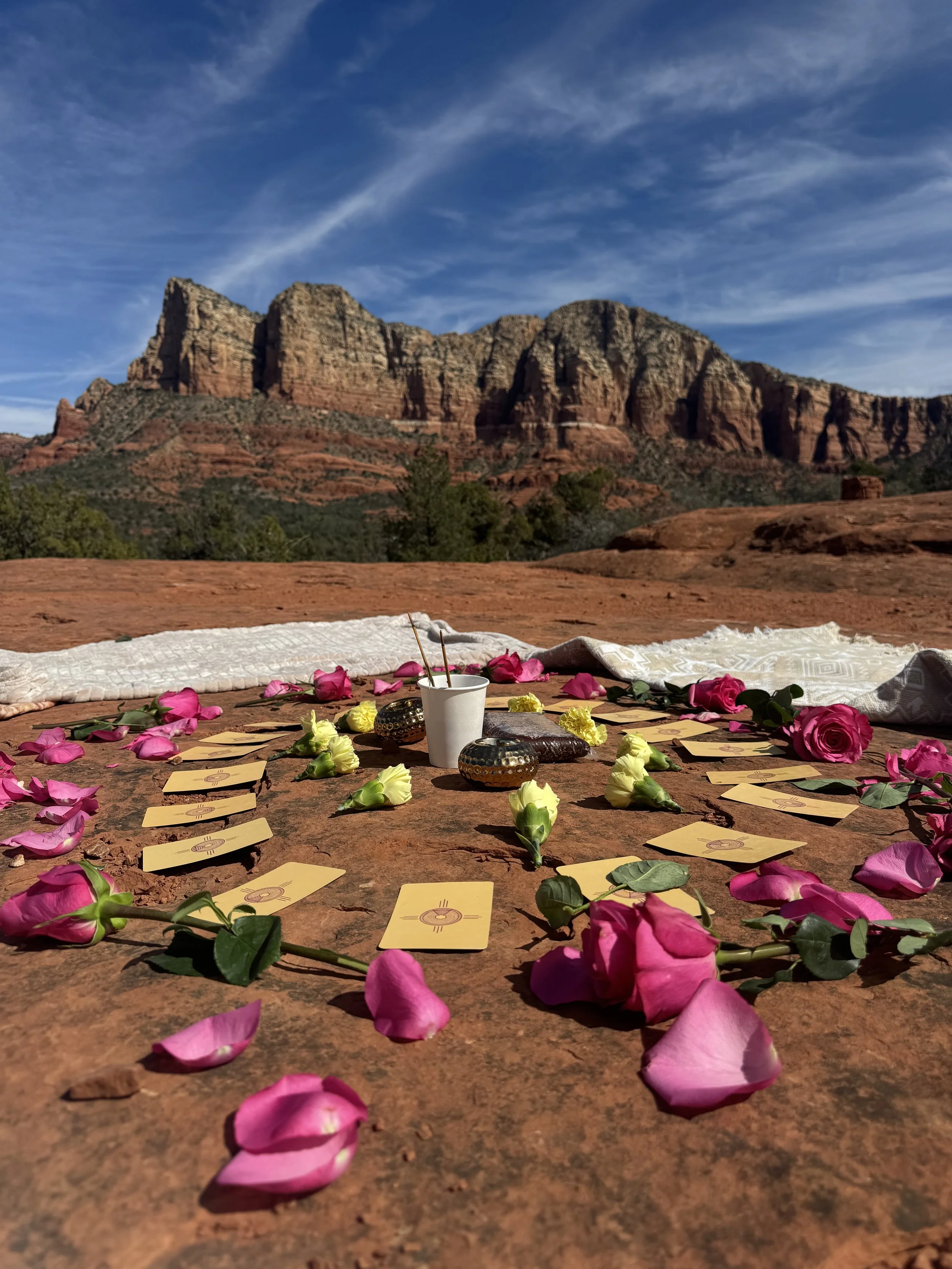 A flat red rock surface with pink and yellow flower petals, tarot cards, a white cup, and decorative objects, with a backdrop of red rock mountains and a blue sky with wispy clouds.