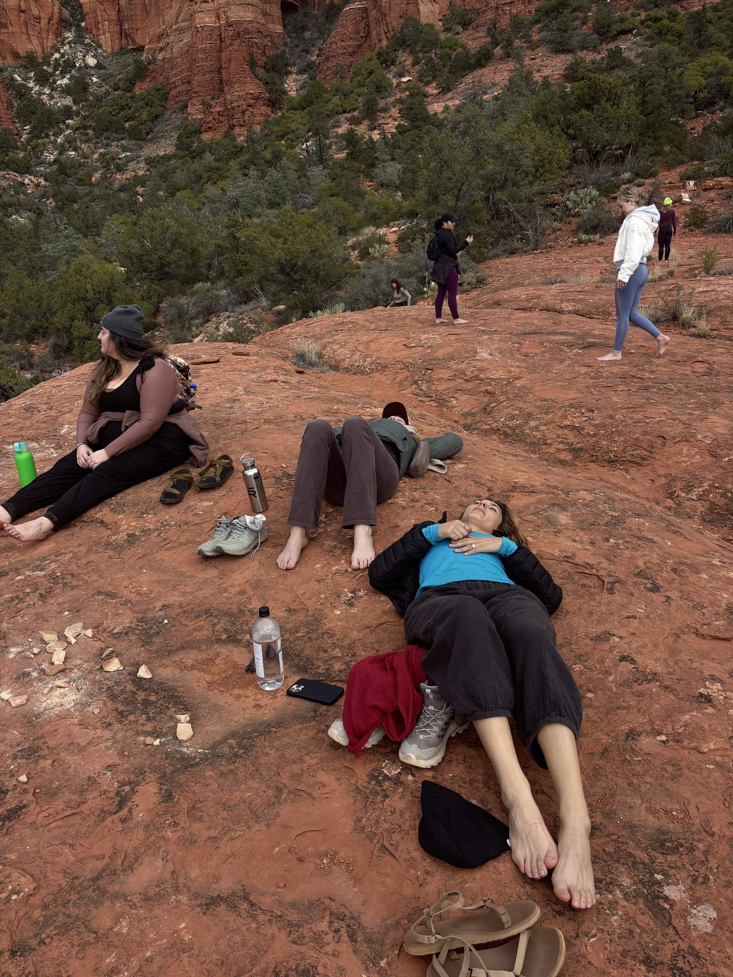 Group of people relaxing and standing on a red rock surface in a desert landscape with sparse vegetation and green trees in the background during daytime.