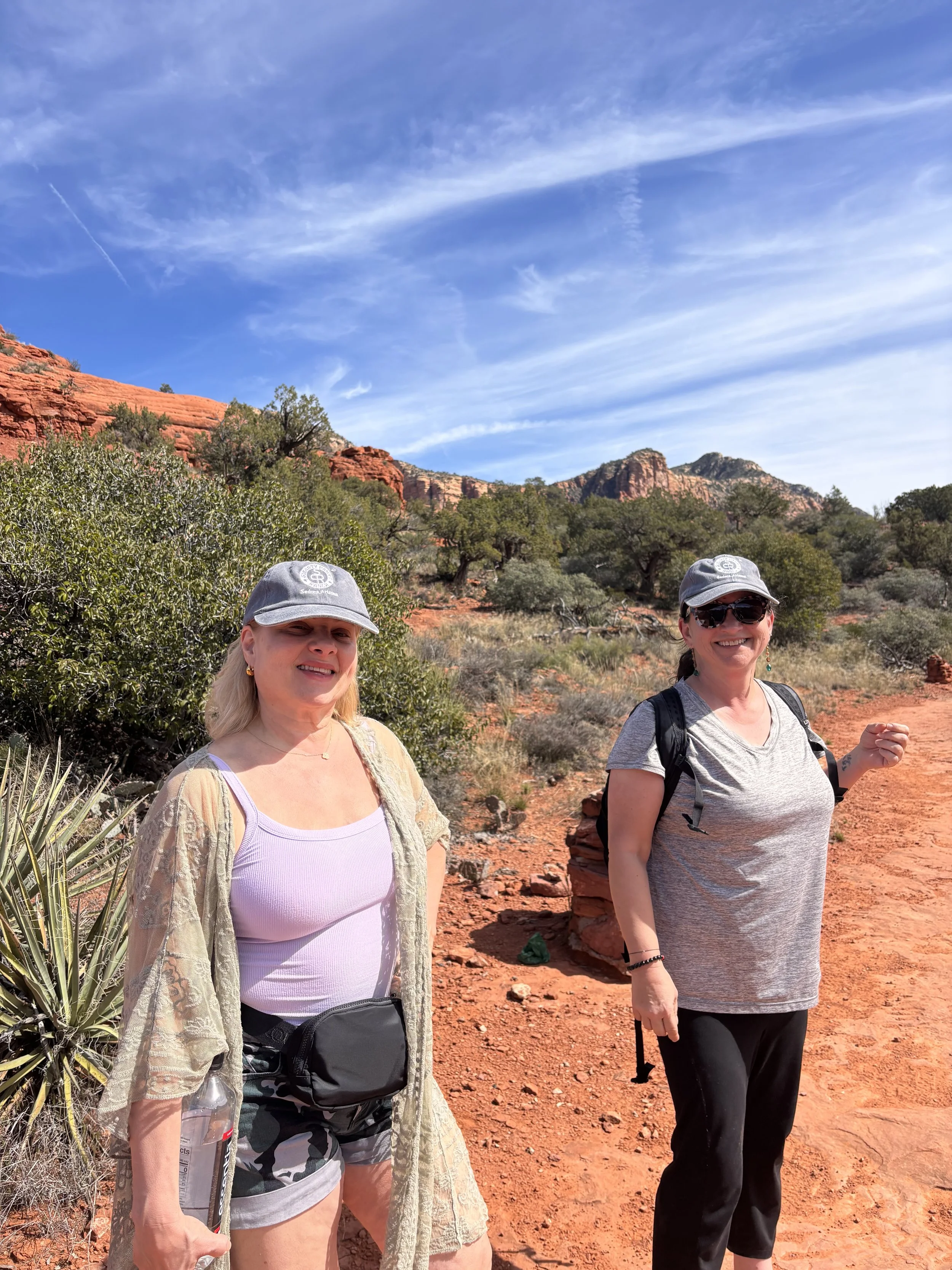 Two women wearing hats and sunglasses hiking in a desert landscape with red rocks and green bushes under a blue sky with wispy clouds.