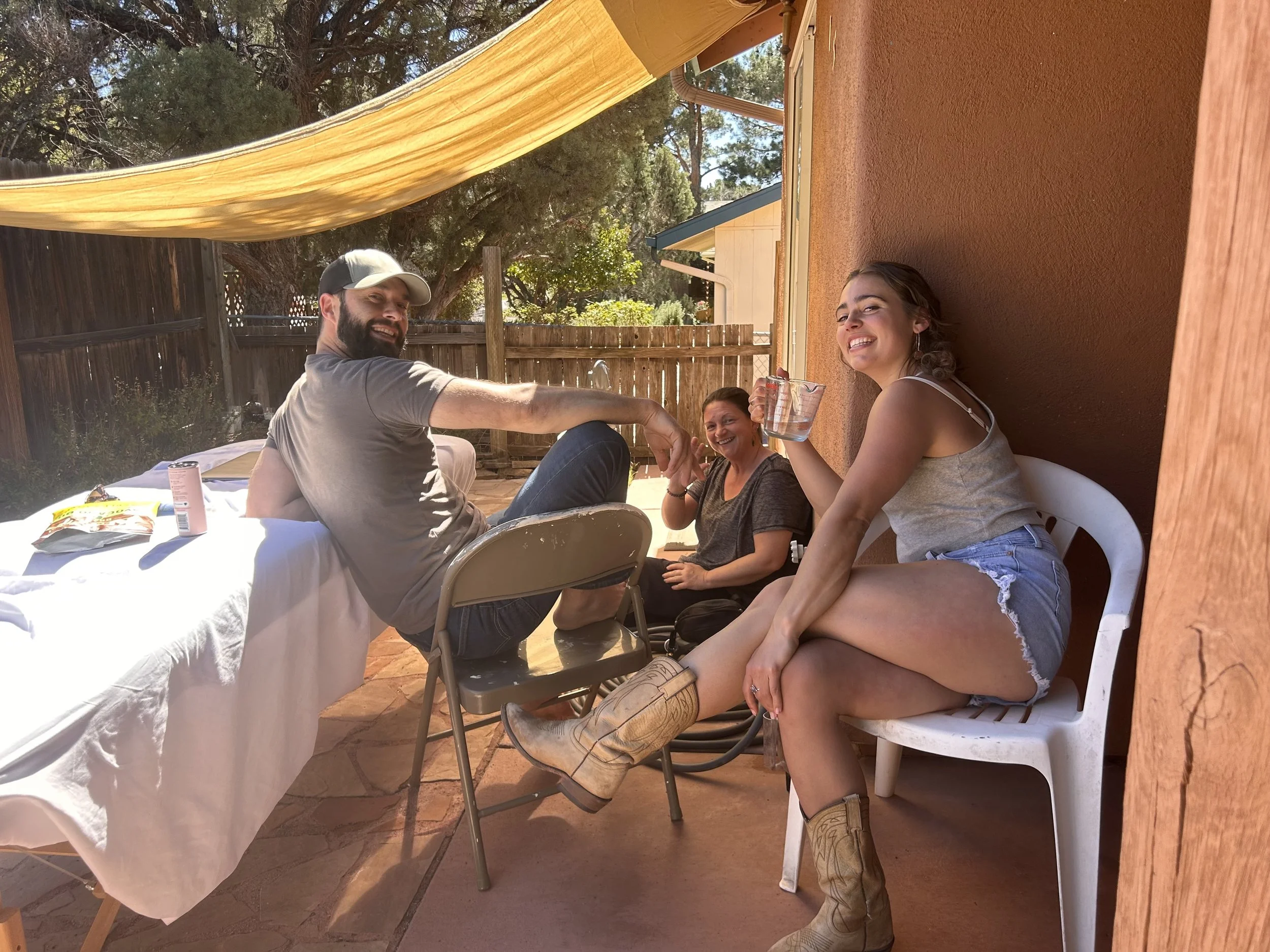 Three people sitting outside at a patio table, smiling and enjoying each other's company. One woman is holding a drink, and another is sitting on a white chair wearing cowboy boots.