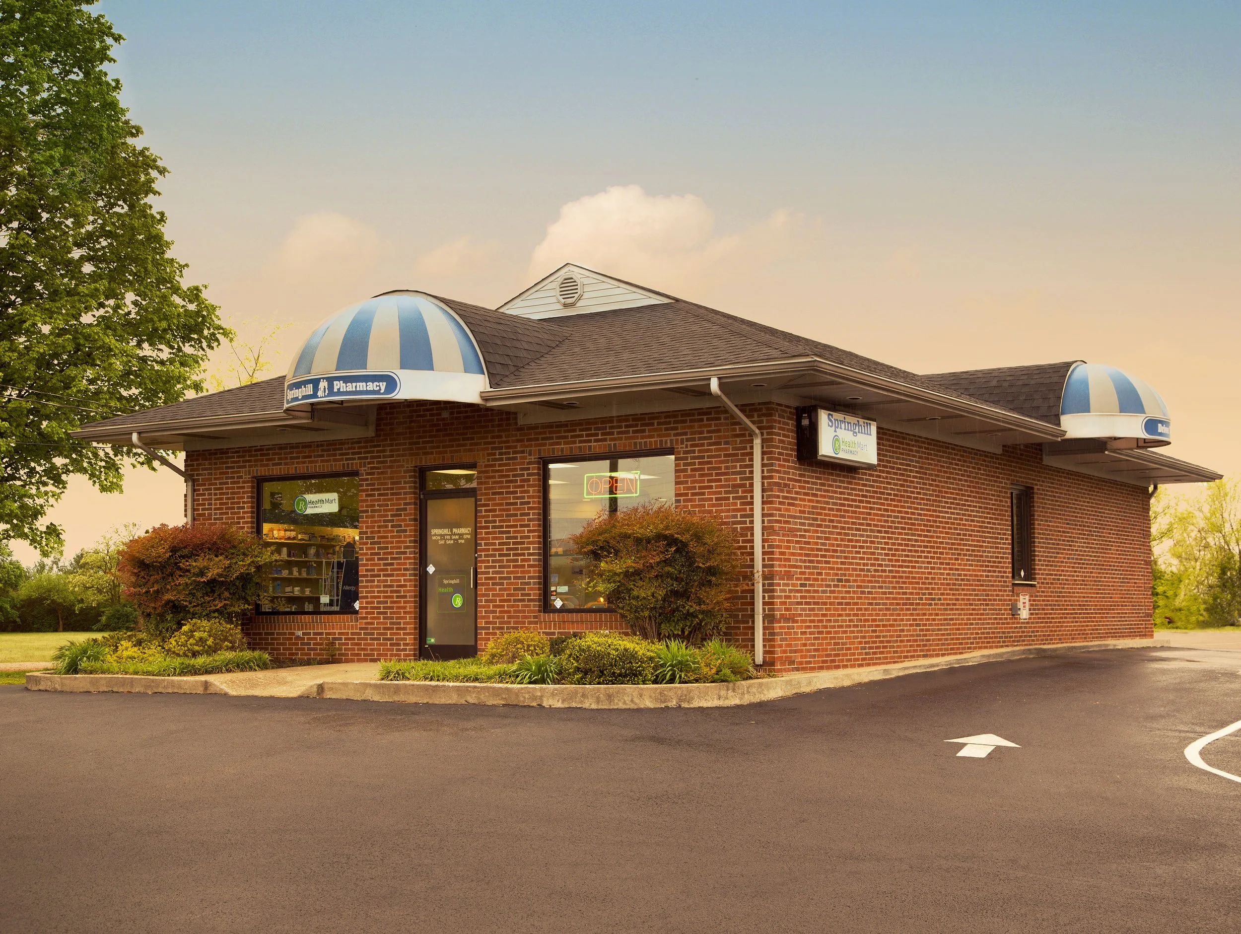A small brick pharmacy building with blue and white striped awnings, a glass door with a neon 'OPEN' sign, and large front windows. The building has a sloped roof, a sign reading 'Springhill Pharmacy,' and is surrounded by a parking lot and greenery, with a sunset sky in the background.