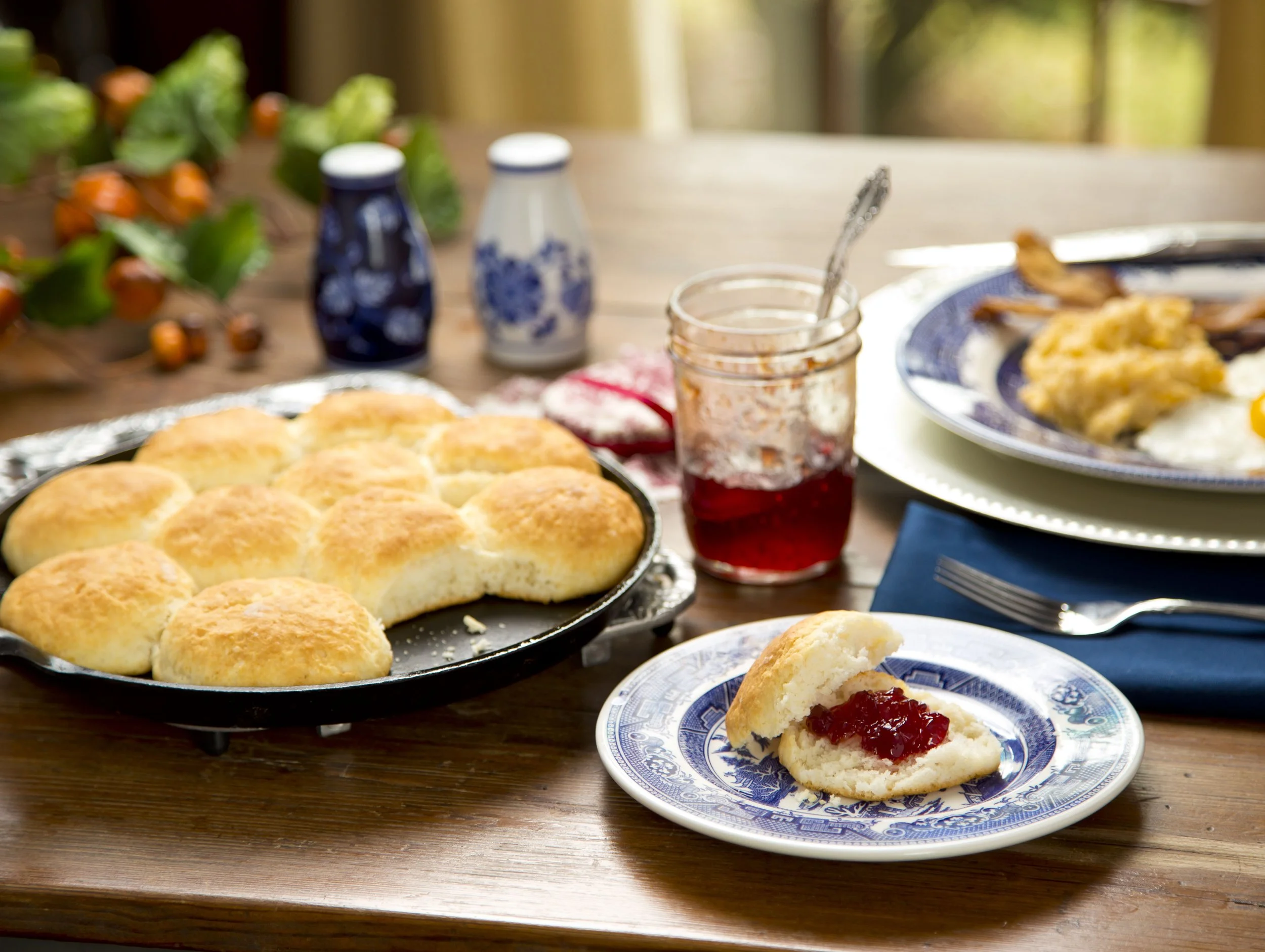 A table set for breakfast with bread rolls, a plate with a biscuit and jam, two bottles of syrup, a jar of jam, a plate with eggs and bacon, and a glass of red juice.