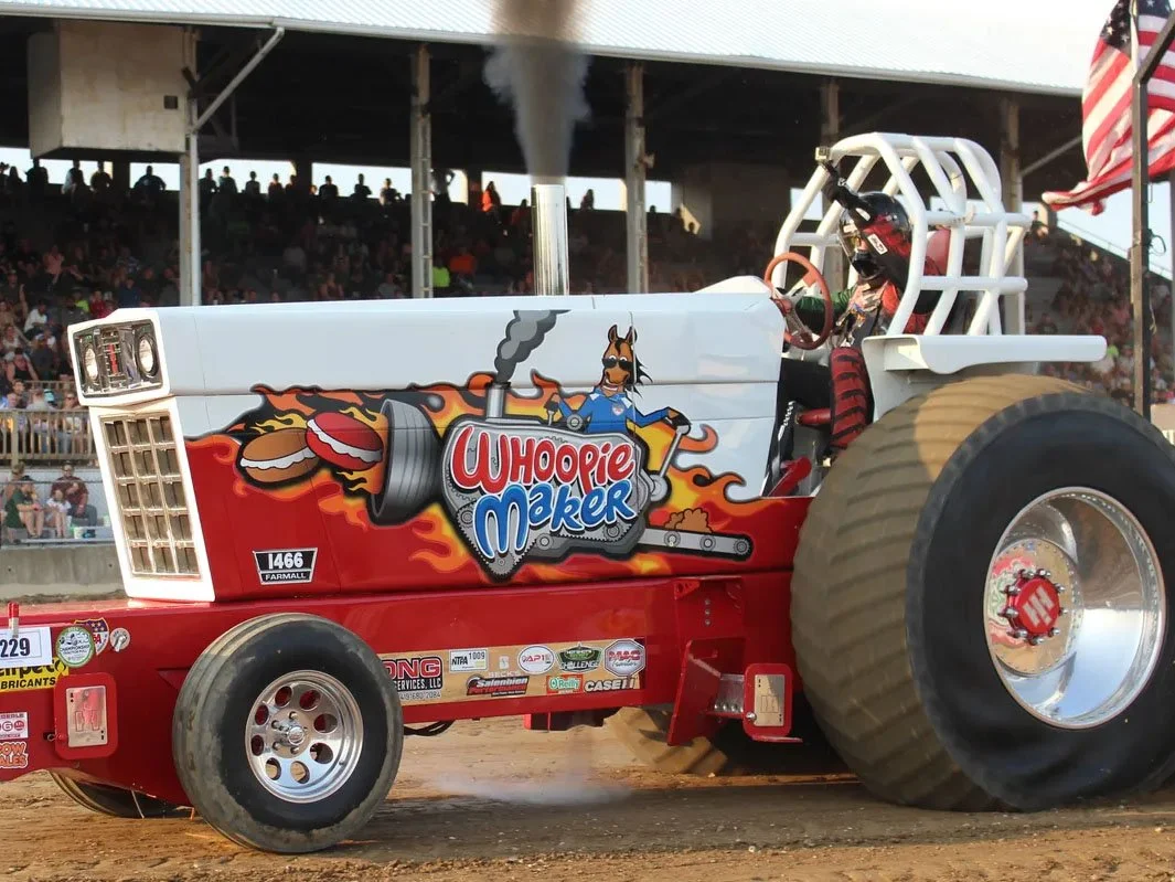 A tractor decorated for a tractor pull with a cartoon image of a donkey, flames, and a Whoopie Maker logo on its side. The tractor is in motion on a dirt track with spectators in the background.