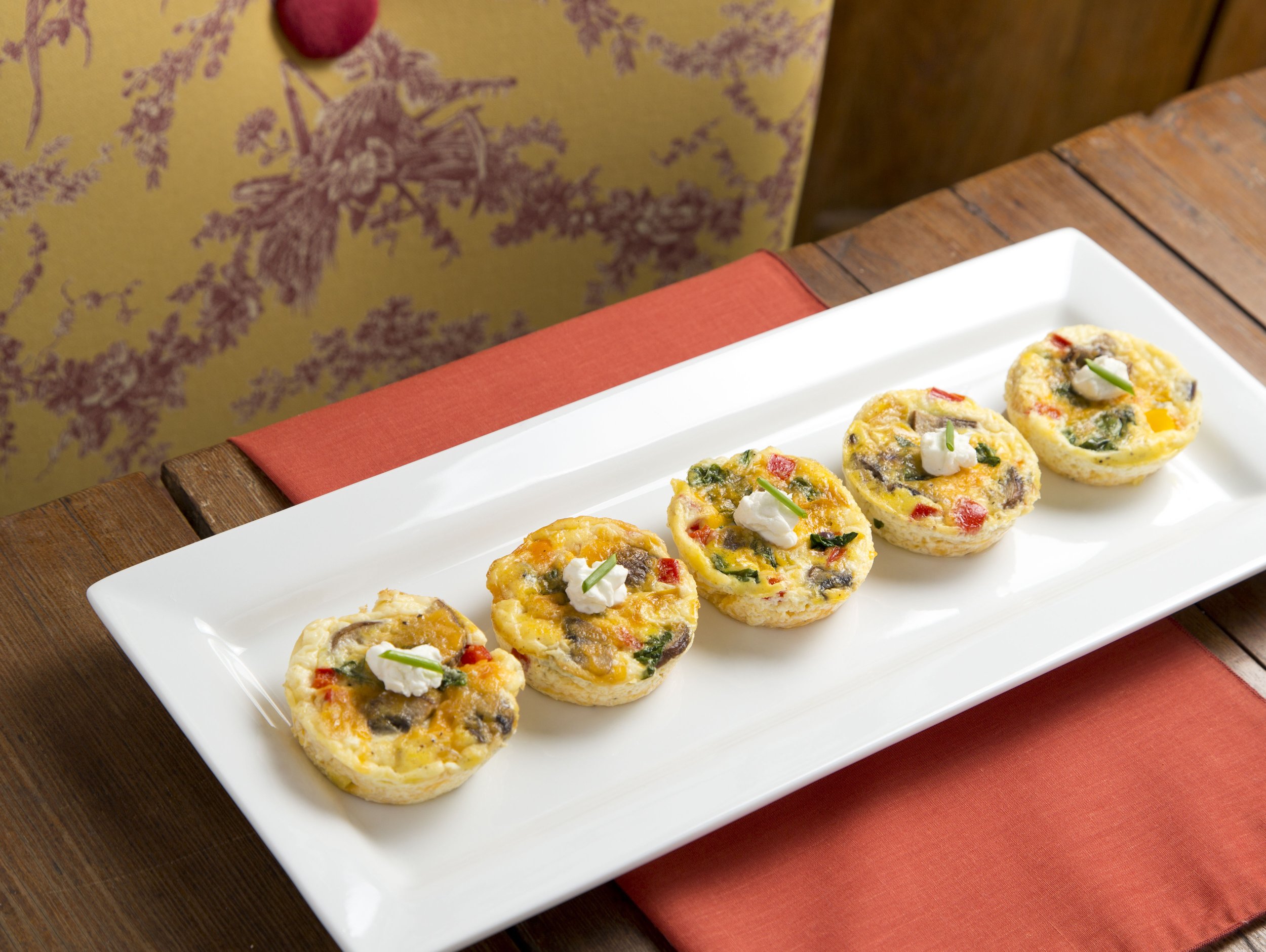 Five mini omelets with vegetables and dollops of sour cream on a white rectangular plate, placed on a wooden table with a red-orange napkin underneath.