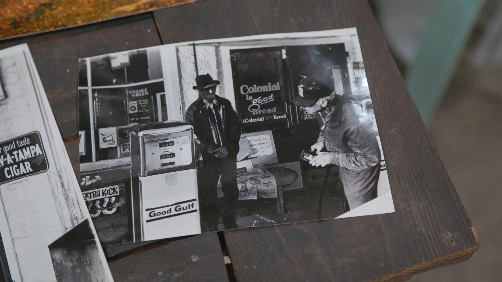 A black and white photo showing two men standing outside a small store with signs that read "Colonial is good Bread" and "Good Gulf". One man is wearing a leather jacket and hat, the other is wearing a cap and smoking a cigarette.
