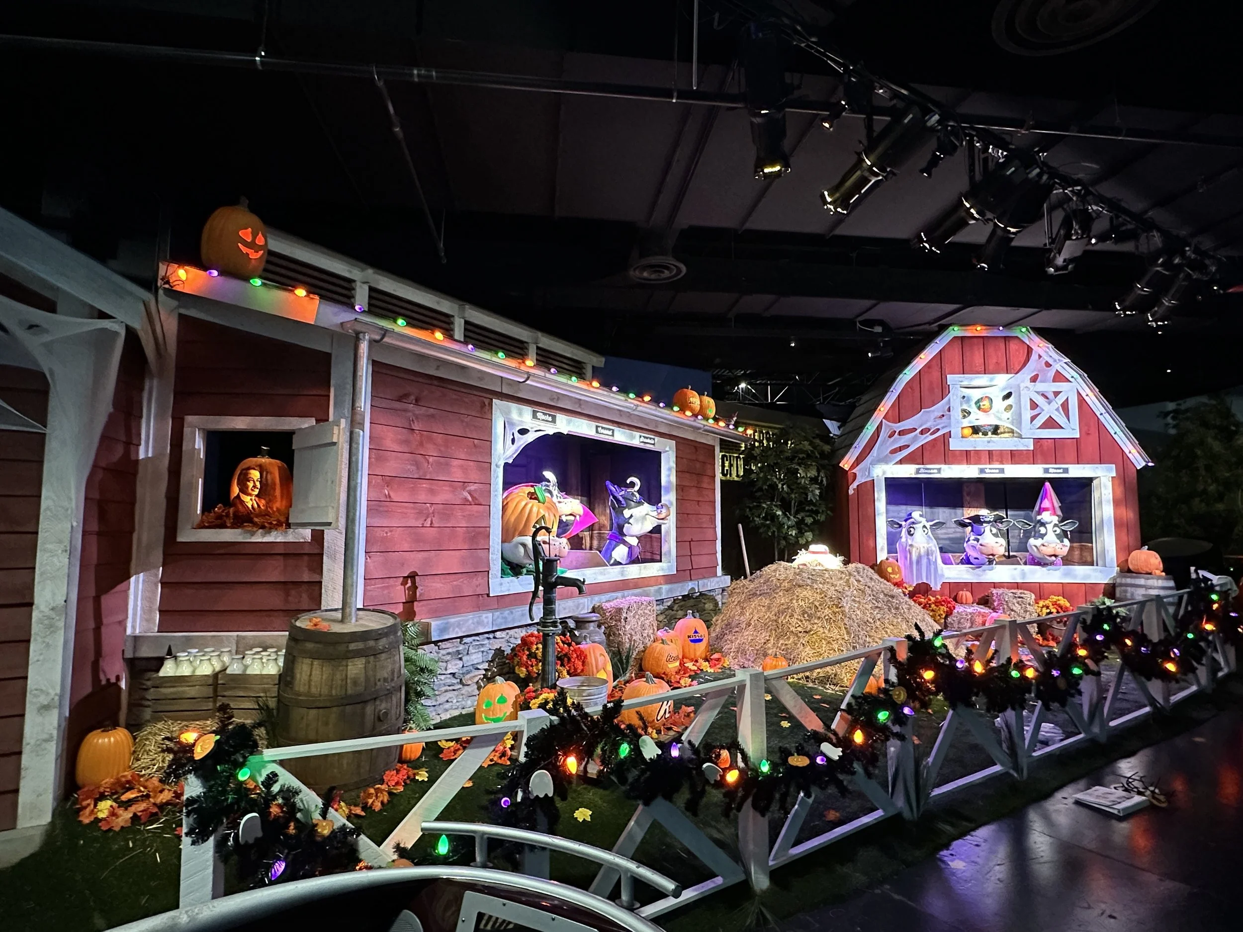 A Halloween display featuring a small red barn with festive decorations, including pumpkins with painted faces, Halloween figurines, cobweb decorations, colorful string lights, and faux hay bales. The scene is lit with ambient lighting for nighttime.