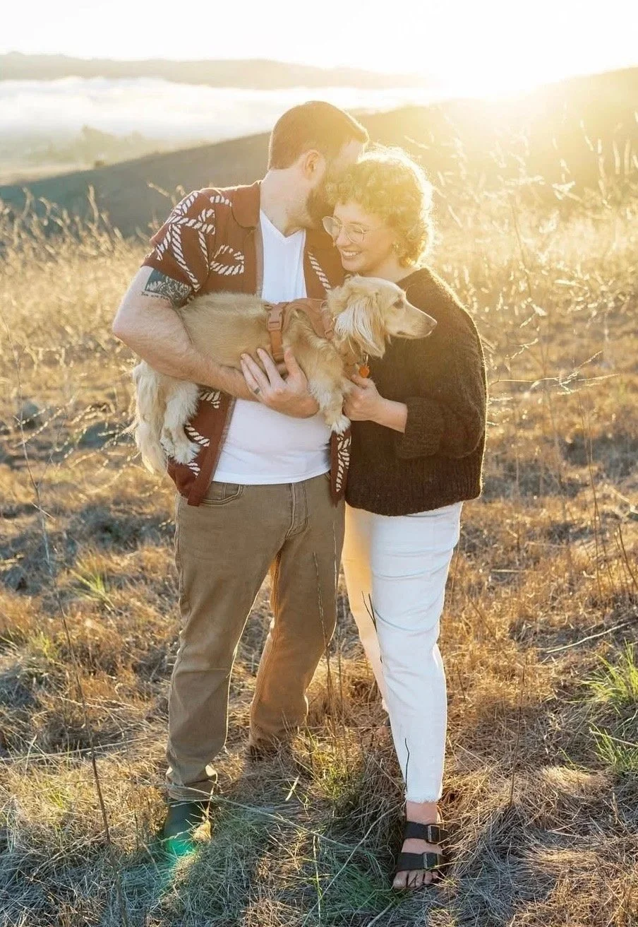 Emma and Lee embrace in a sun-kissed field in the PNW with their dachshund in their arms.
