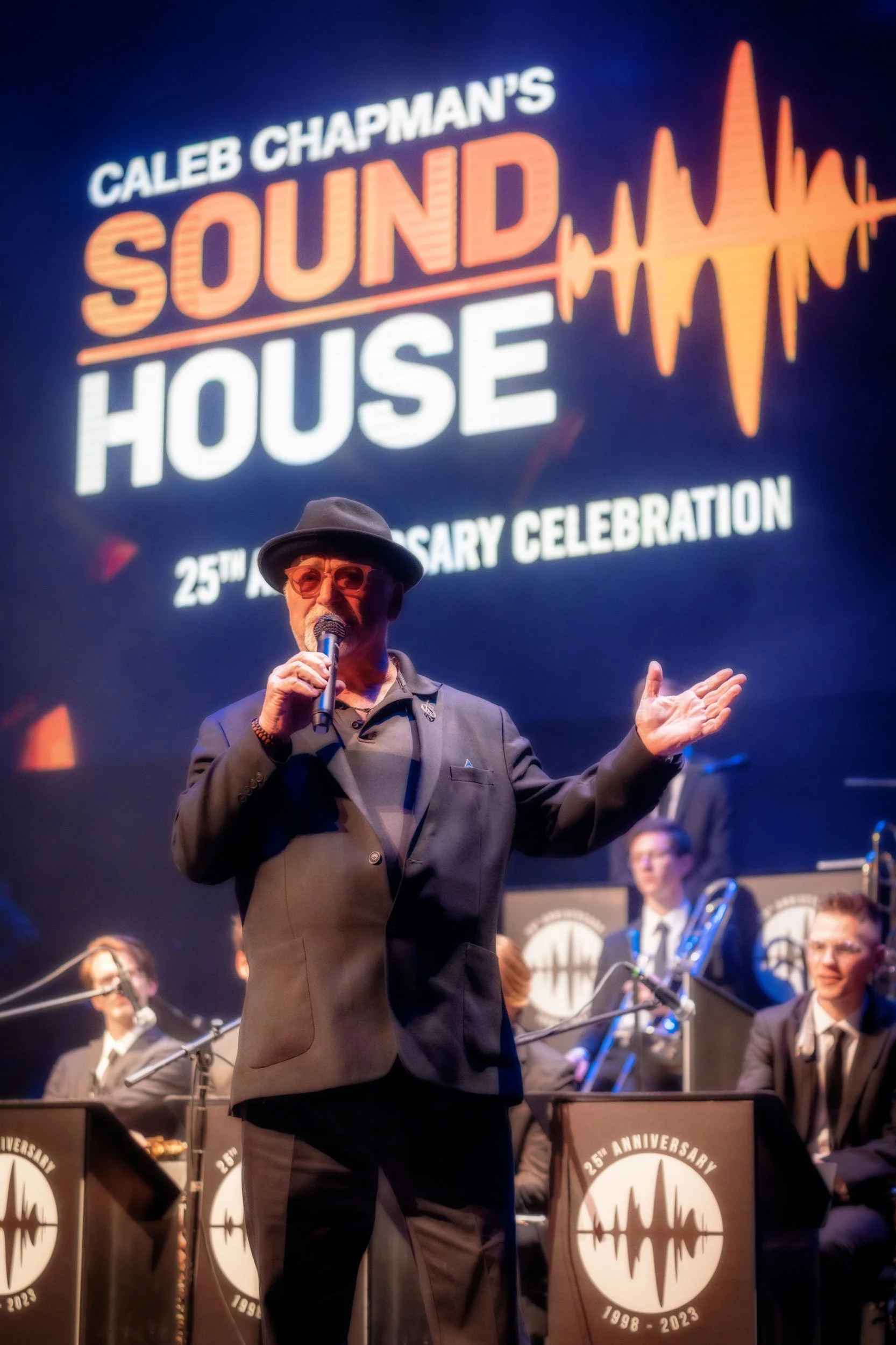 Person in black suit and hat speaking into a microphone at "Caleb Chapman's Sound House" 25th anniversary event, with musicians and logo in the background.