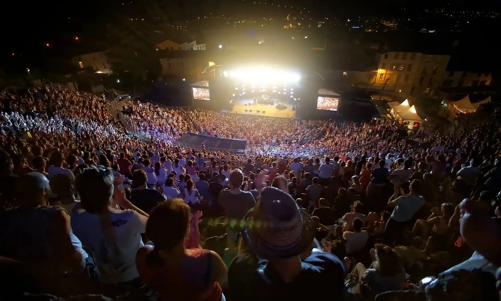Amphitheater crowded with people attending an outdoor concert at night, with bright stage lights illuminating the stage.