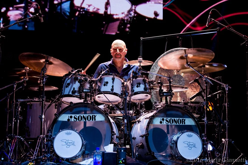 A drummer playing a Sonor drum set on stage with cymbals and microphones around.