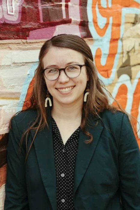 A woman with glasses and earrings smiling in front of a colorful graffiti wall.