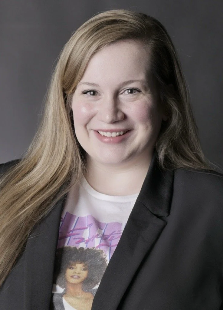 A smiling woman with long blonde hair, wearing a black blazer over a graphic T-shirt, posing against a gray background.