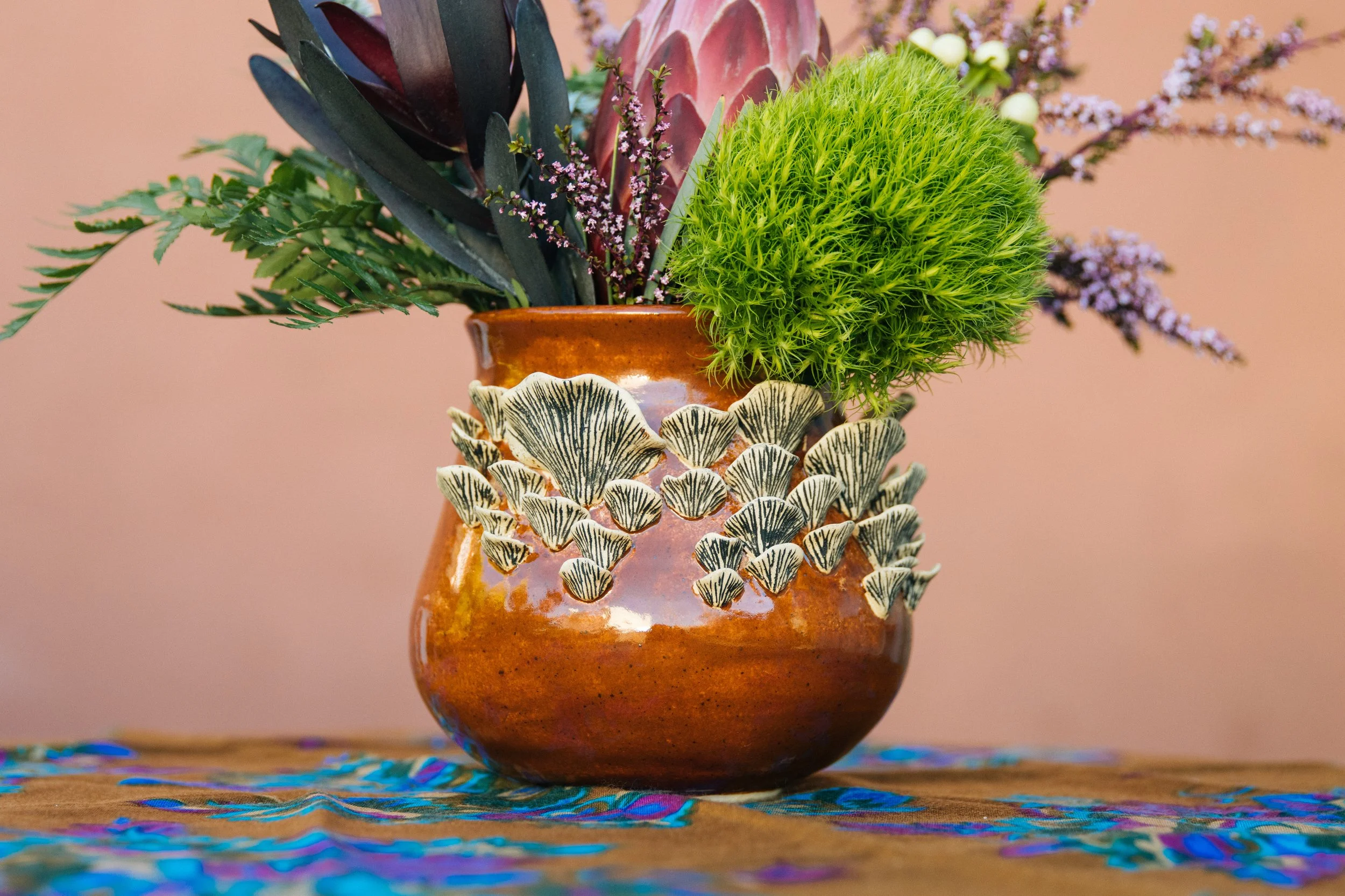A decorative ceramic vase with black and white patterned mushrooms attached to it, filled with various green foliage and purple flowers, sitting on a colorful cloth surface.