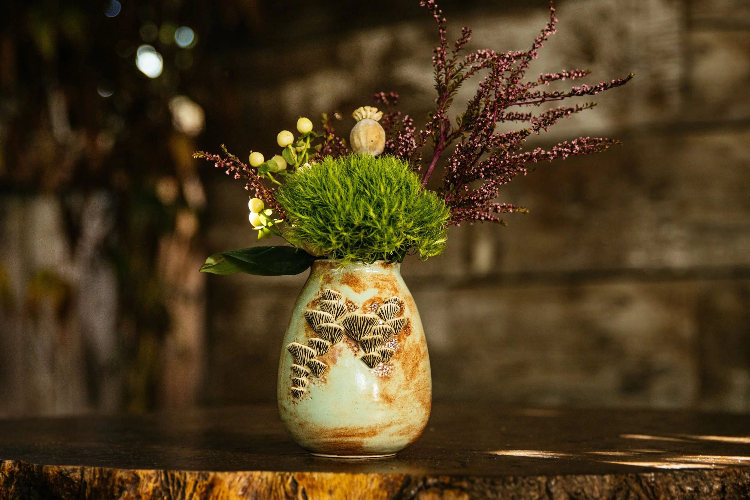 A ceramic vase with a decorative pattern of black and beige mushrooms, containing an arrangement of green moss, purple and white flowers, and small yellow berries, set on a wooden surface with a blurred wooden background.