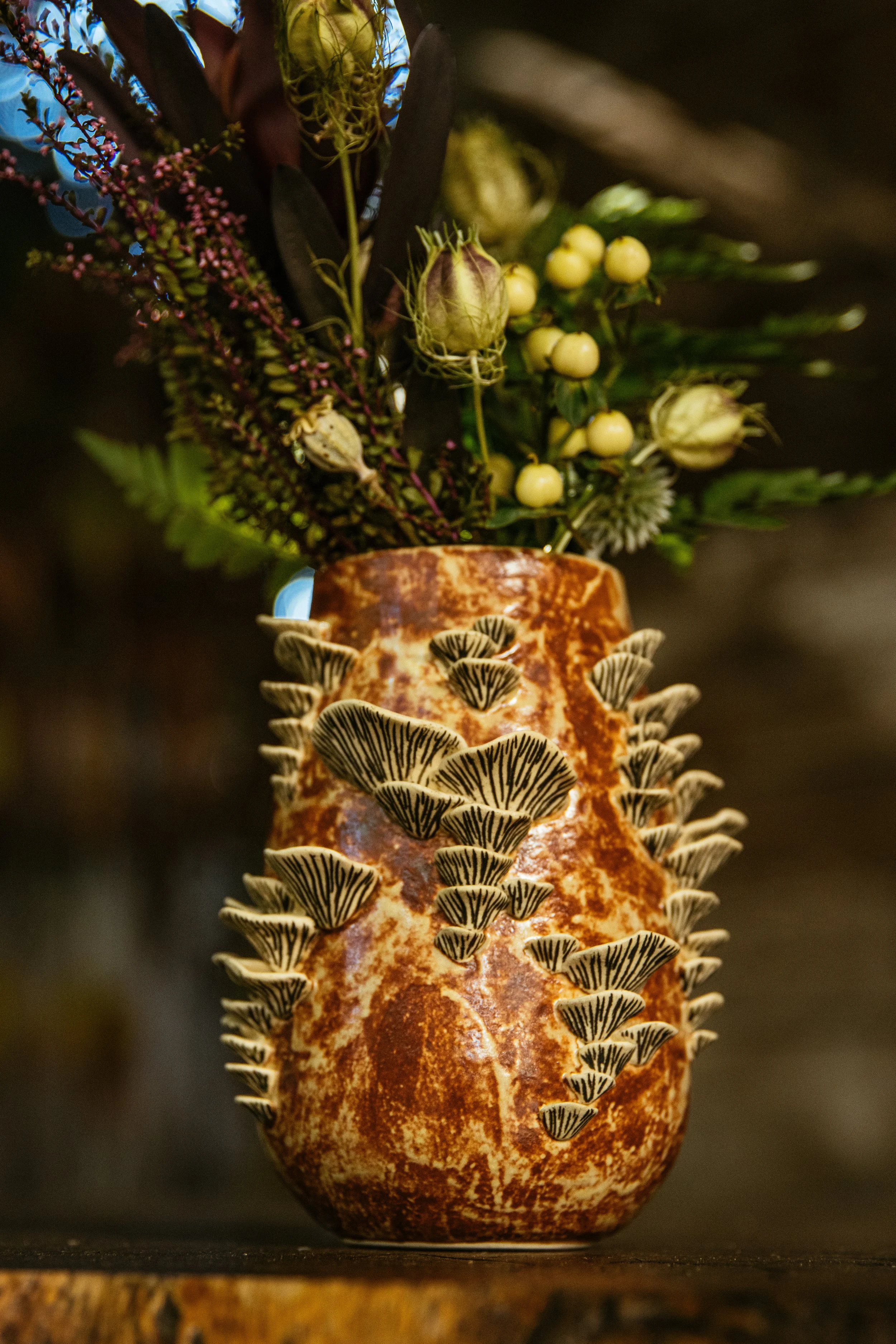 A ceramic vase with a rust-colored glaze and protruding black-and-white striped mushrooms, filled with flowers and plants, placed on a surface.
