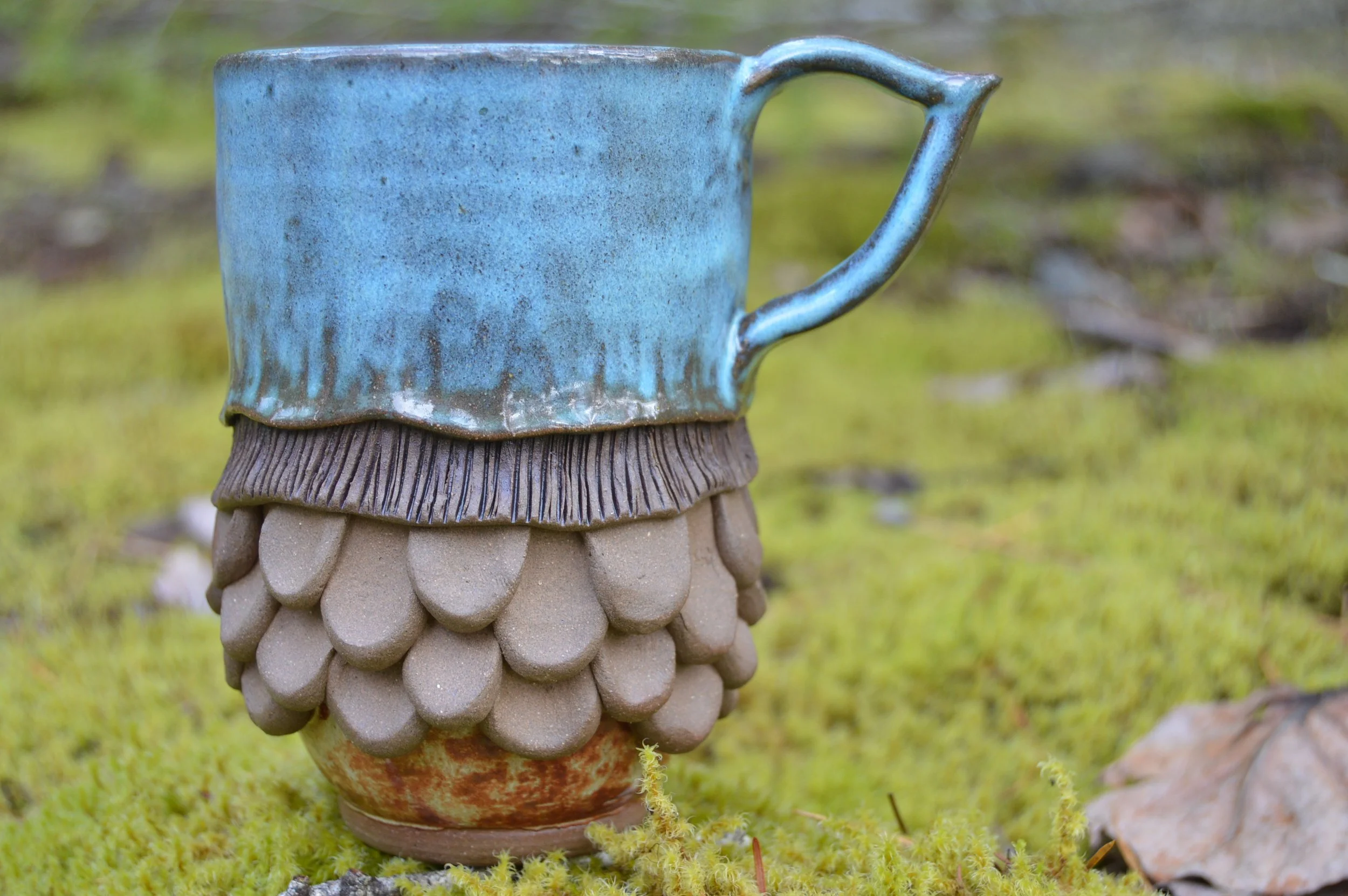 A whimsical and elaborately sculpted ceramic mug with a blue glaze on the upper part, a textured brown middle section resembling mycelium, and a base resembling a pinecone, placed on mossy ground outdoors.