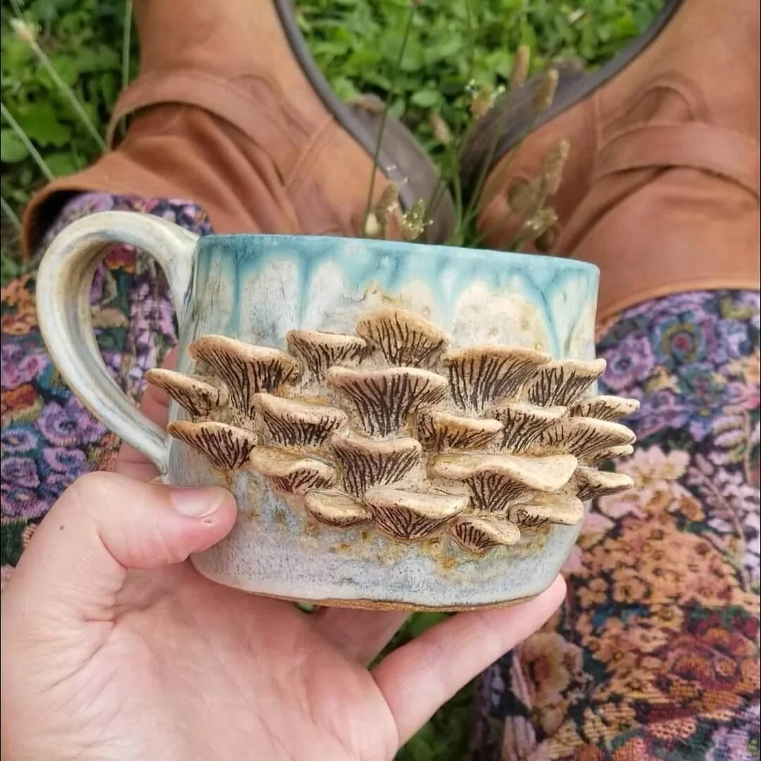 A person holding a ceramic mug decorated with mushroom reliefs, with grassy ground and brown boots in the background.