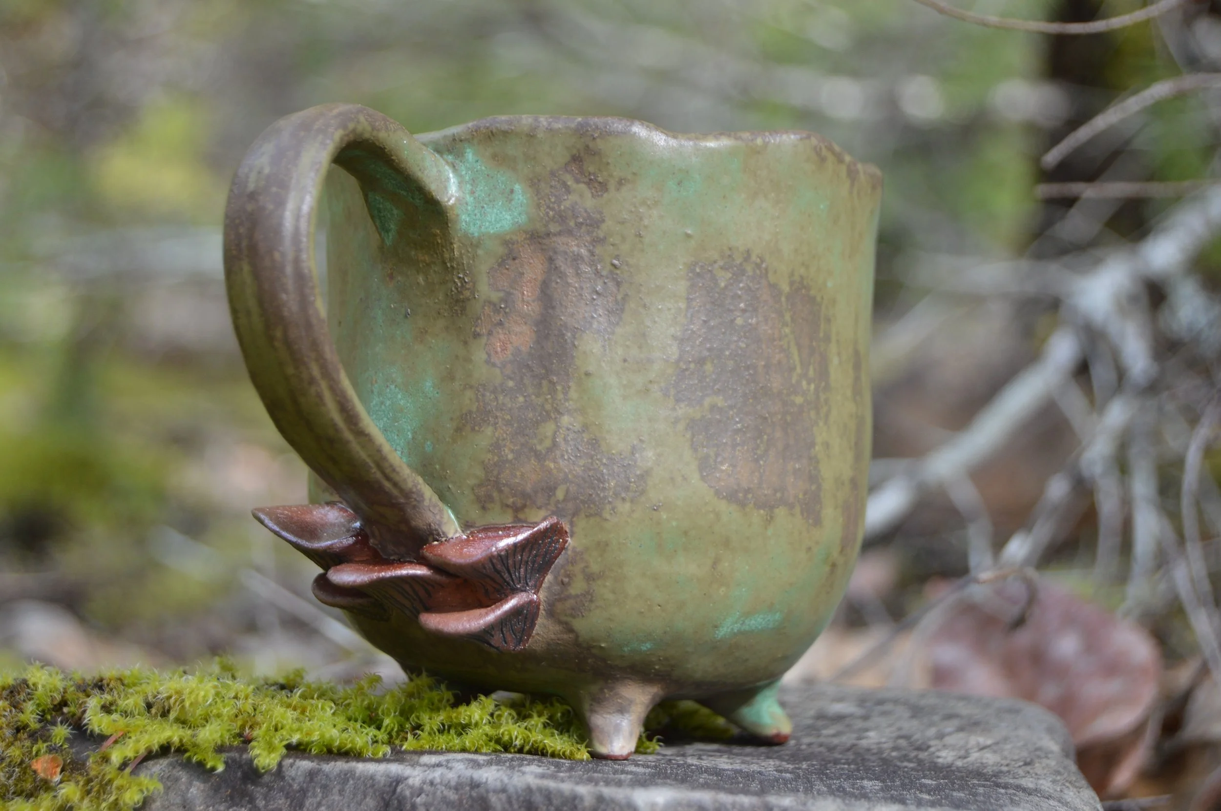 A ceramic mug with a cactus-shaped handle and three small feet, sitting on a rock with moss and surrounded by foliage.