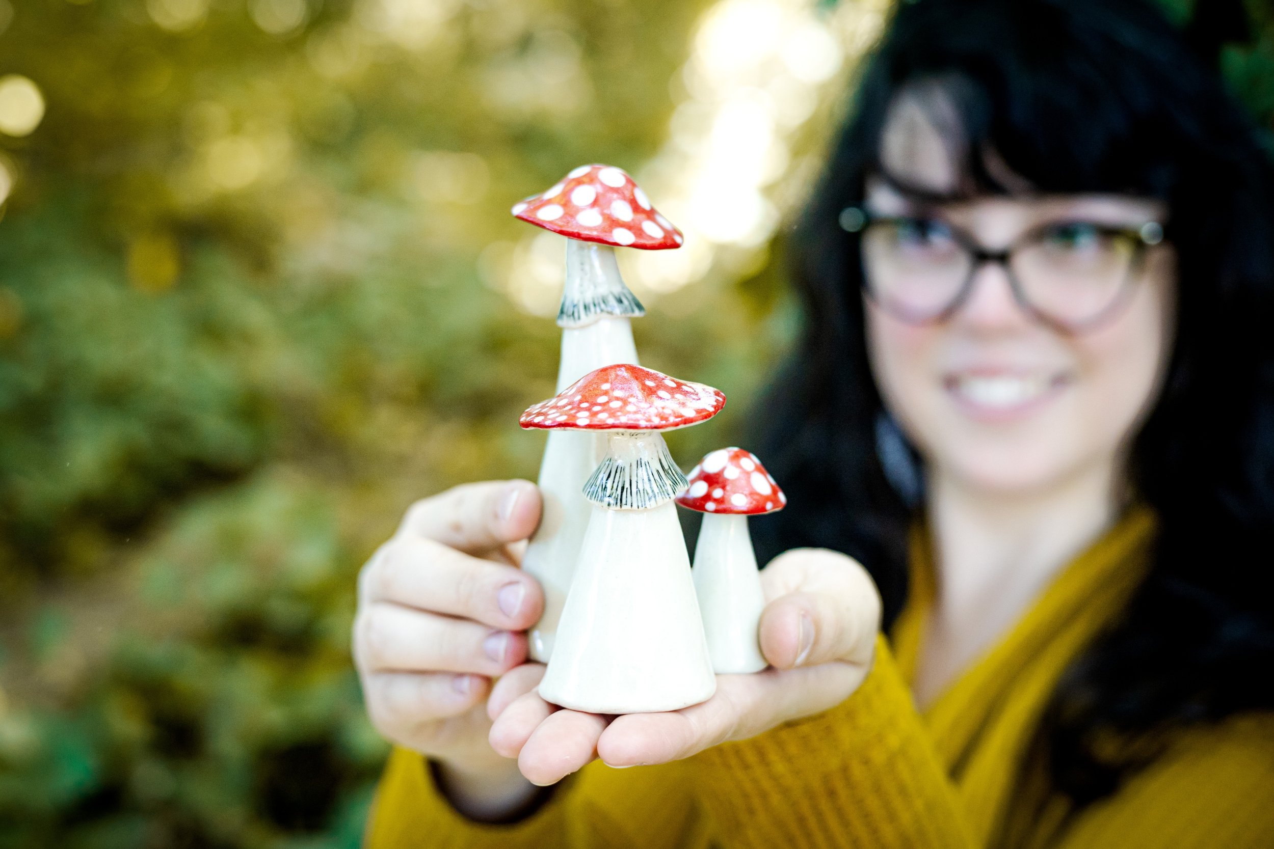 Jenna Vannest pictures with black hair and glasses, holding colorful ceramic Amanita Mushroom sculptures.