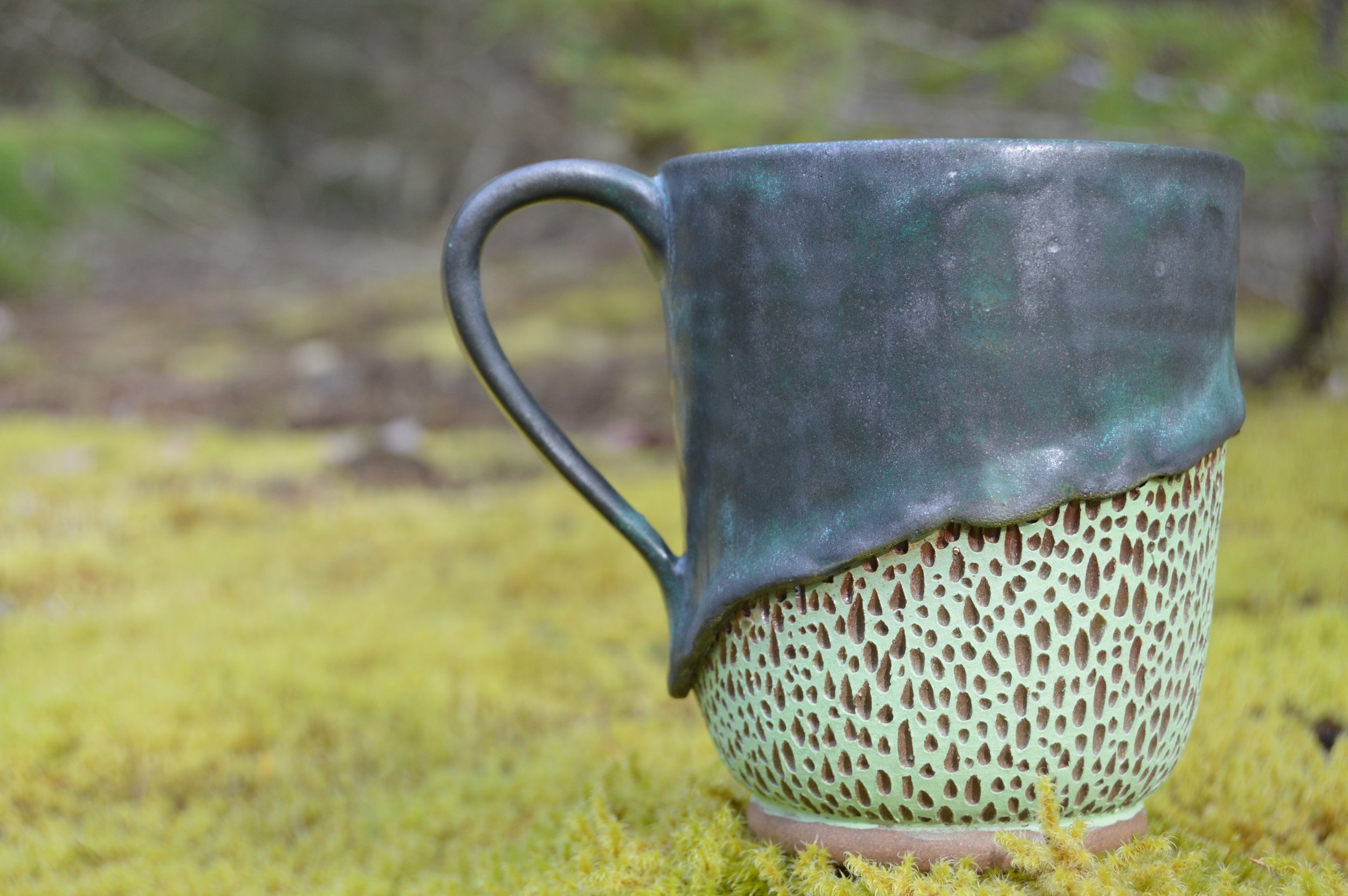 A ceramic mug with a dark greenish glaze on top and an intricate, open lattice pattern on the lower half, placed on a mossy surface.
