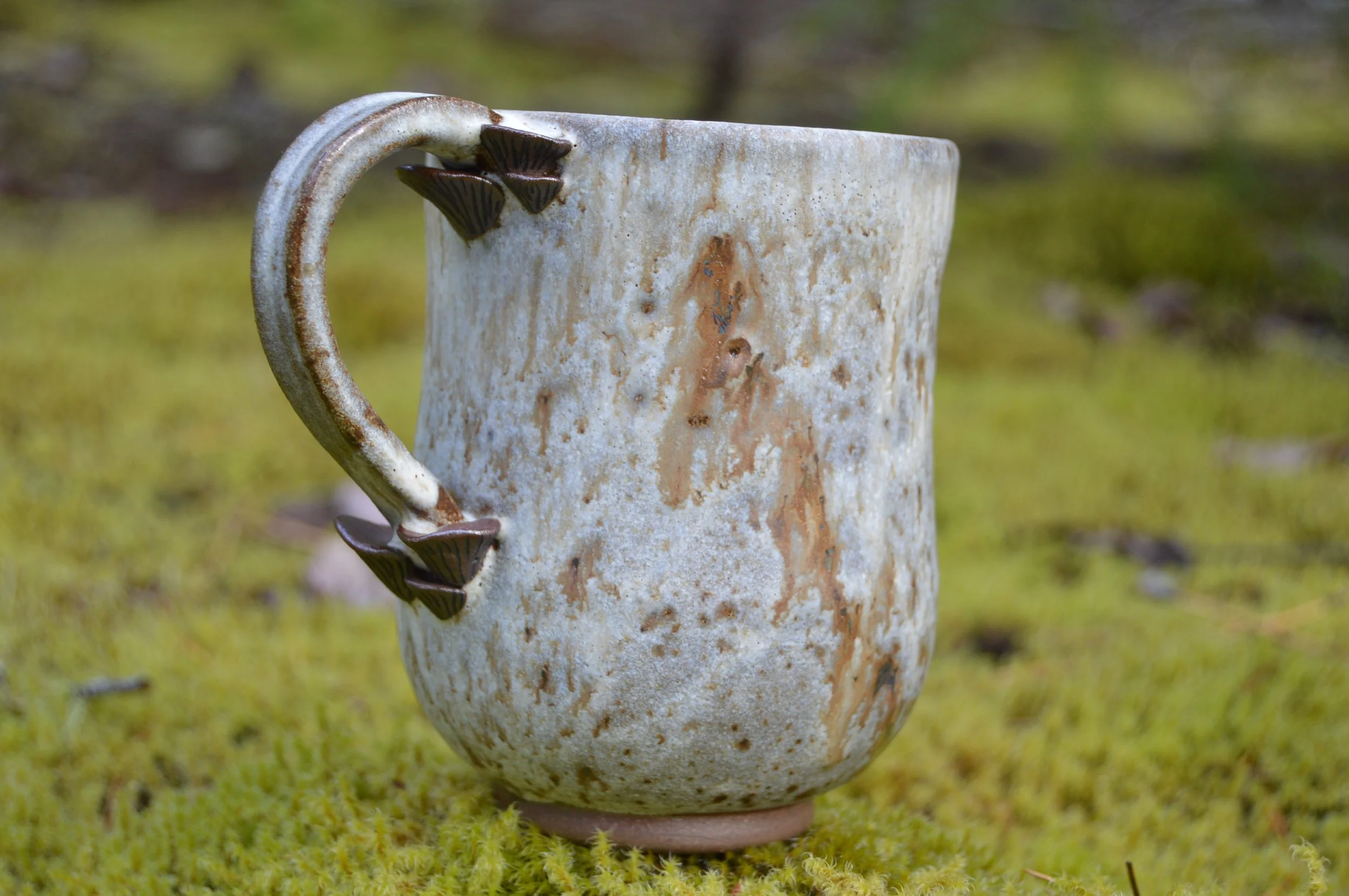 A ceramic mug with a rustic, weathered appearance, sitting on mossy ground outdoors. The mug has a decorative handle with carved details shaped like mushrooms.