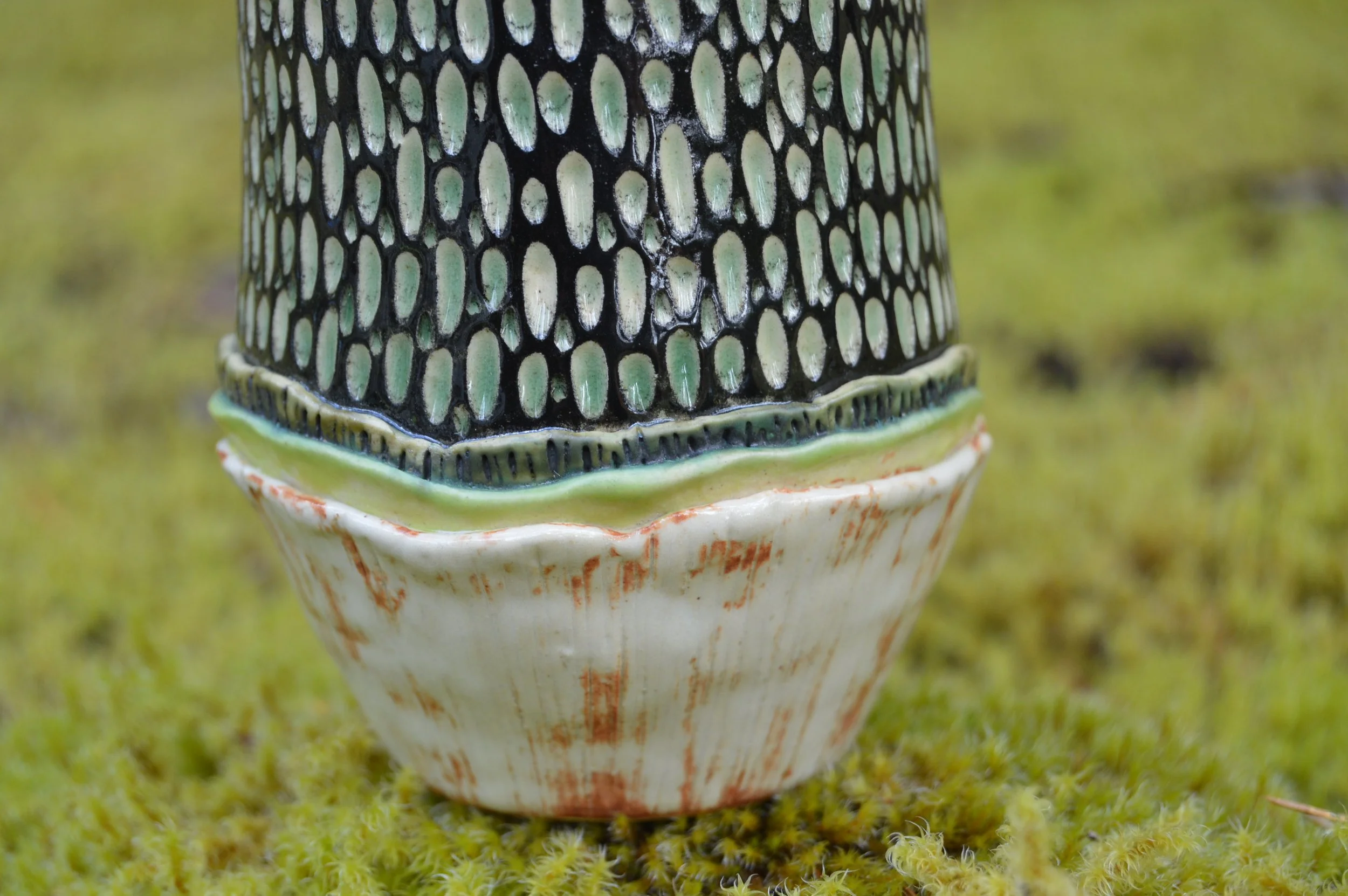 Close-up of decorative ceramic object with a black and green textured pattern on top, resting on a scalloped white base with reddish-brown accents, on a mossy green surface.