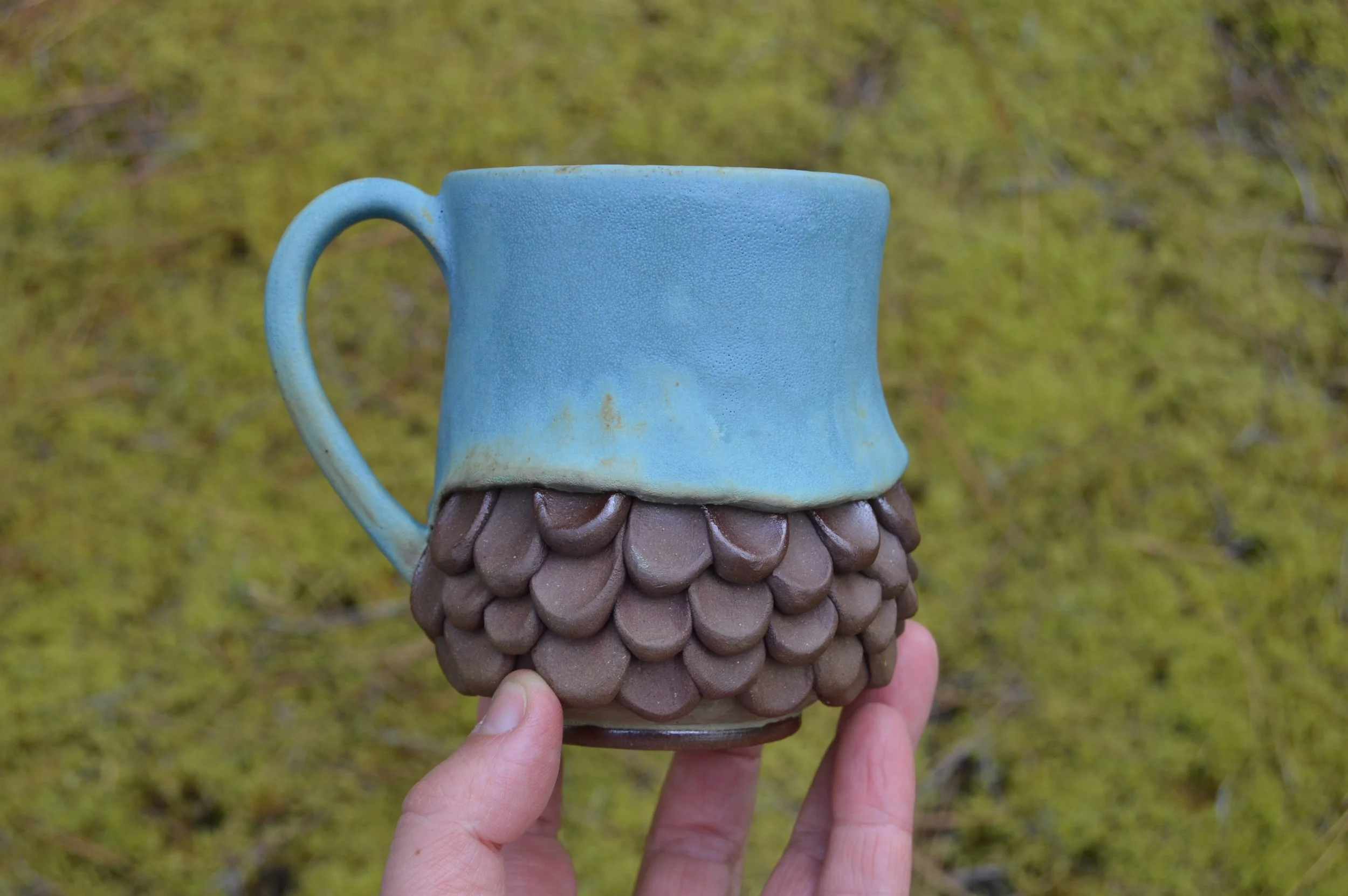 A ceramic mug with a blue upper half and a brown textured lower half resembling fish scales, held by a person's hand against a blurred green background.
