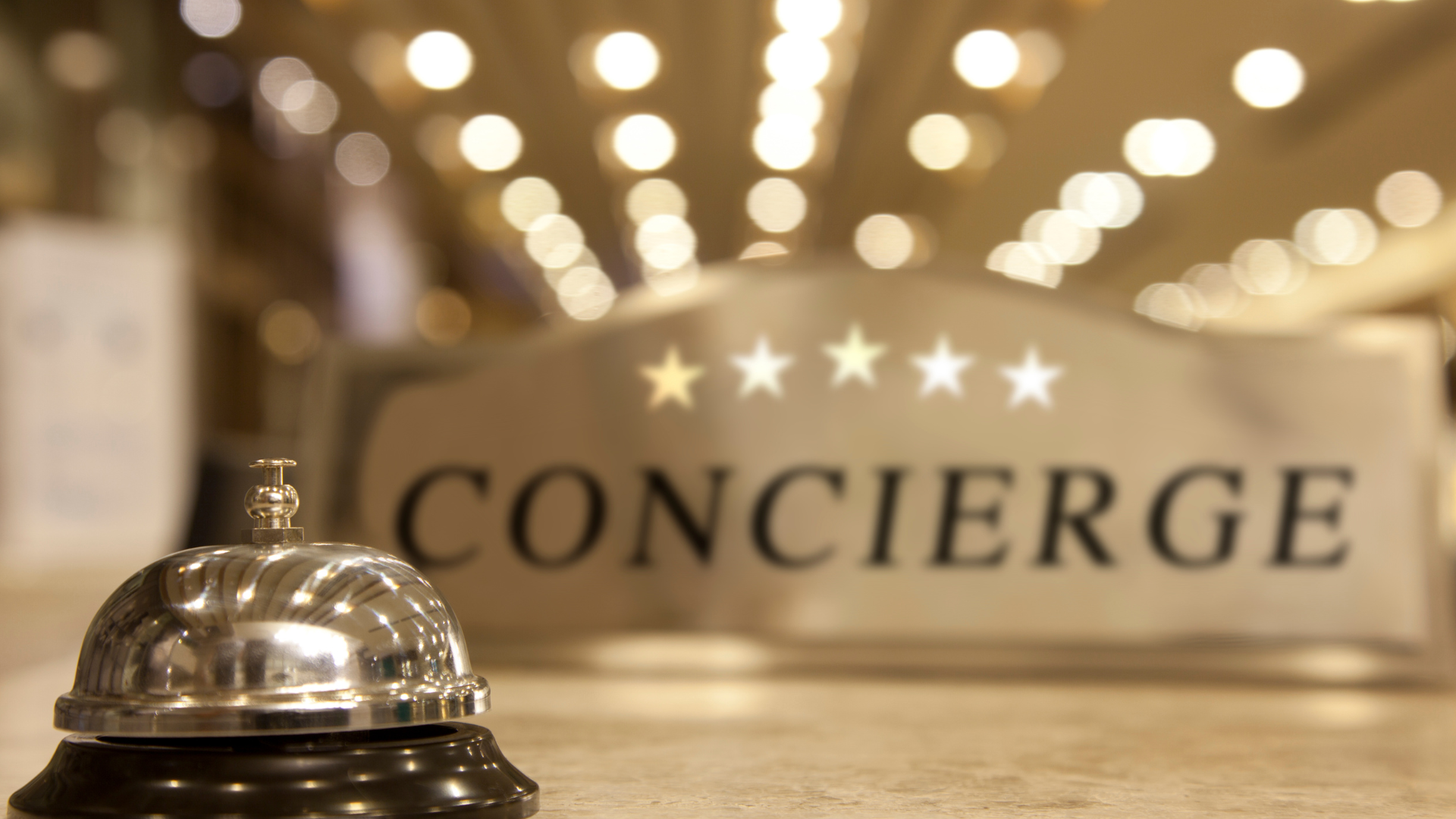 A shiny bronze bell sits on a white surface with a blurred background of a metal sign that reads 'Concierge' and has five white stars above the text. There are also warm, out-of-focus lights in the background.