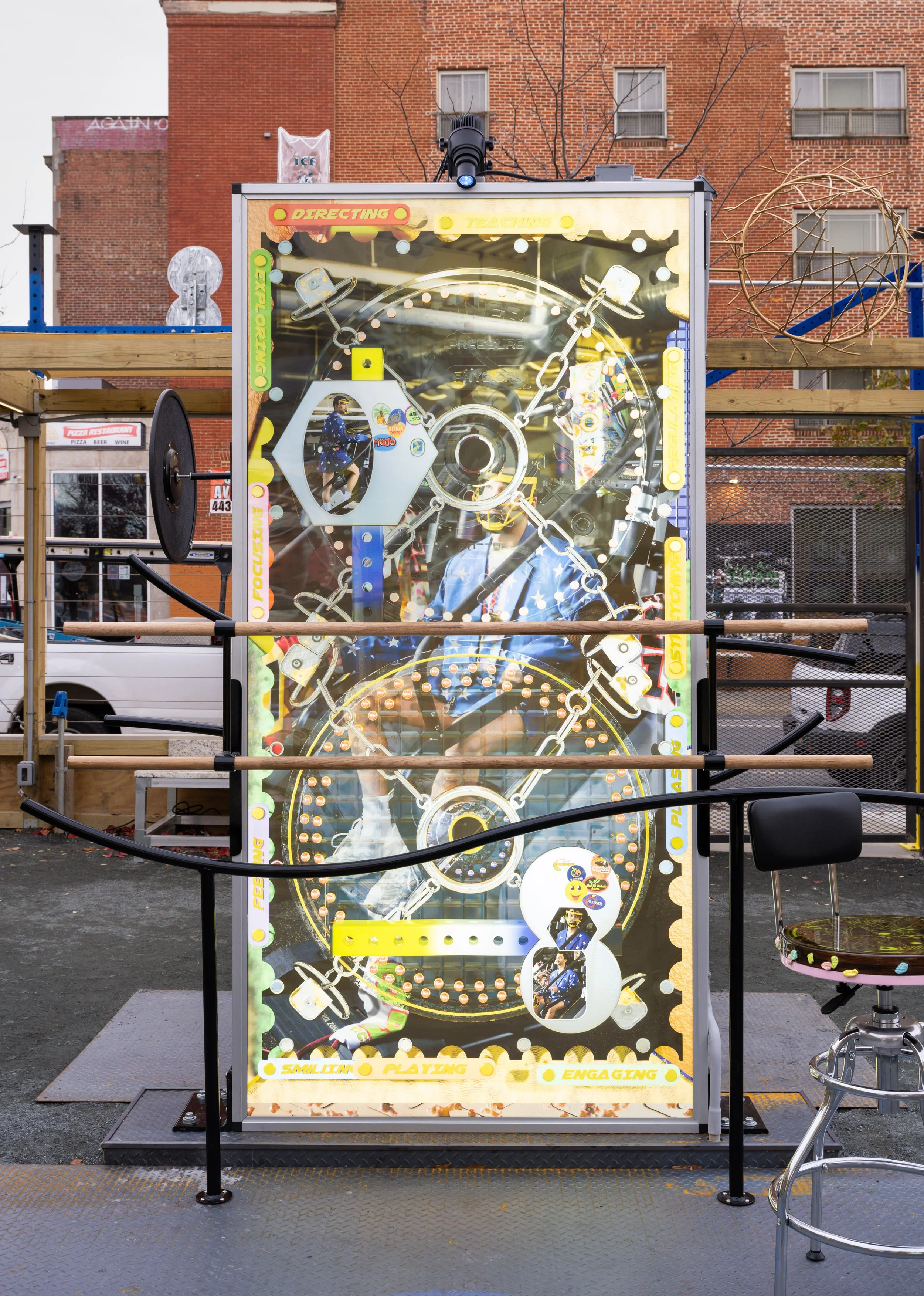 An art instillation with the theme of engaging and playing, decorated with colorful stickers and lights, situated outdoors on a black metal platform with barriers around it.