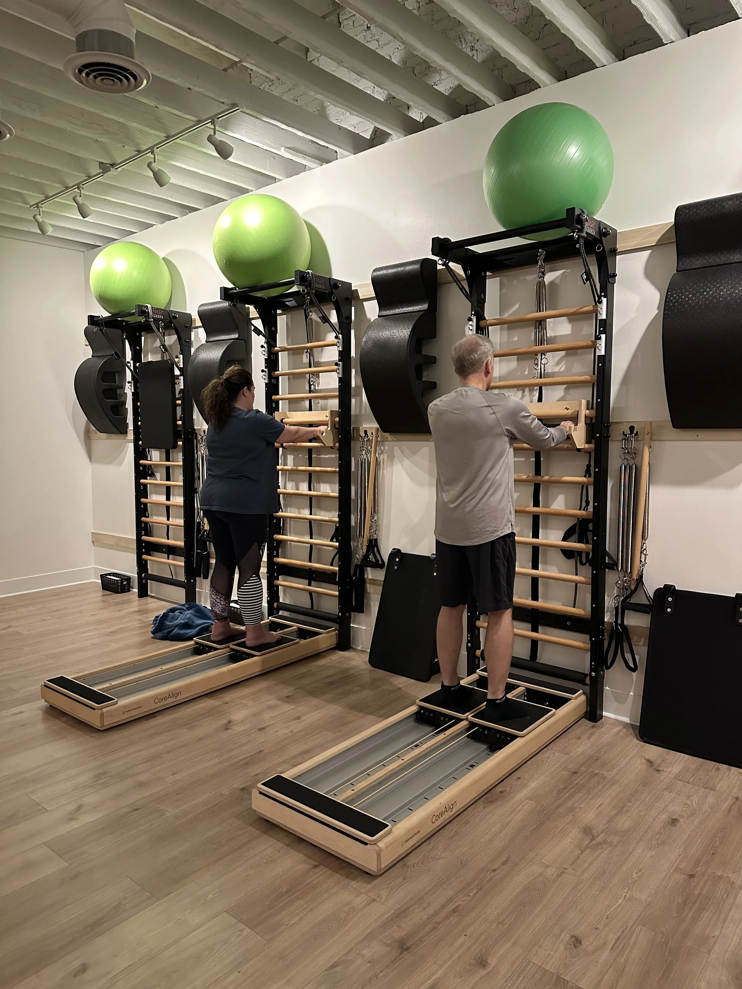 Two people using Pilates reformer machines in a gym, with green exercise balls mounted on the wall above.